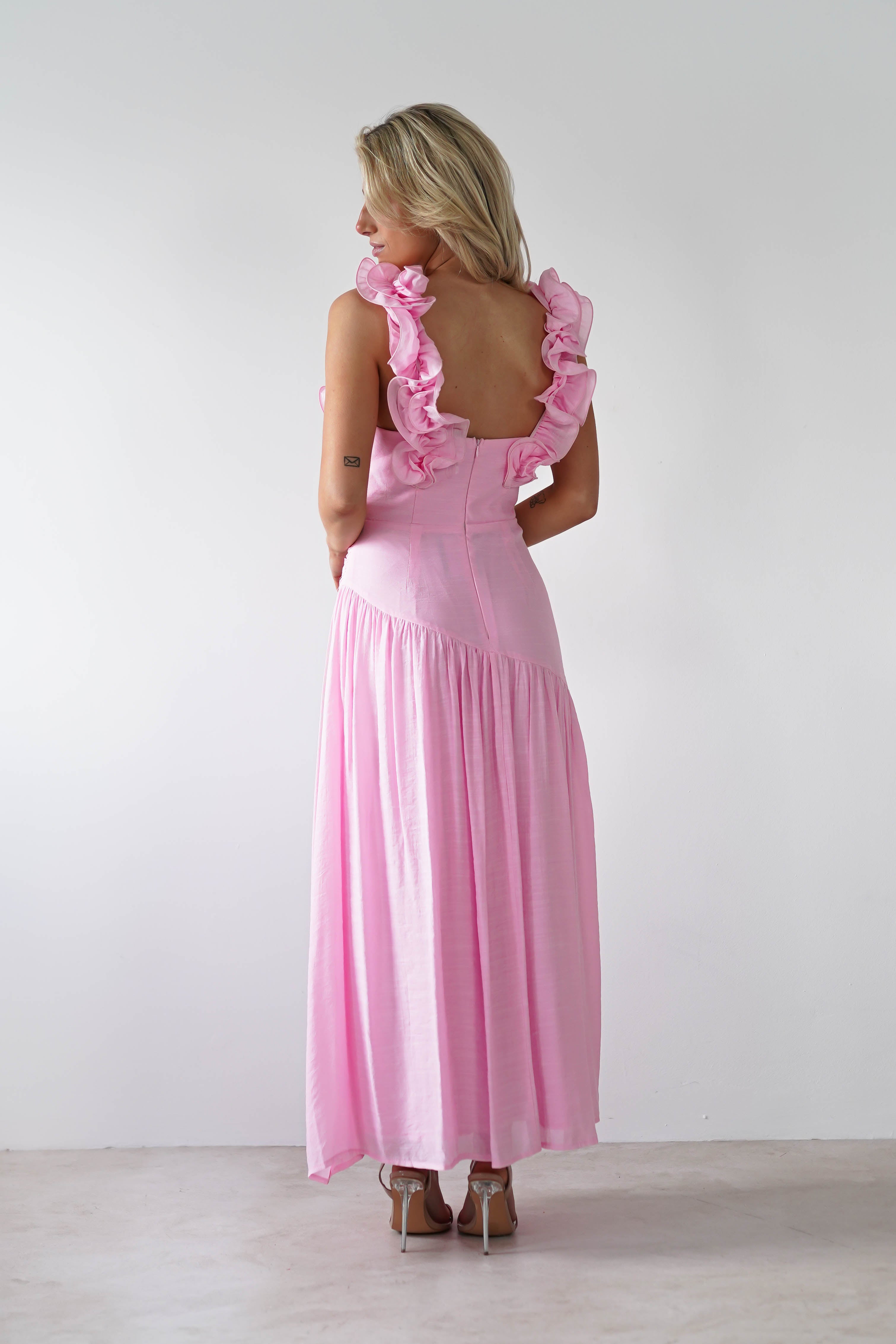 A woman with blonde hair stands in a neutral-toned room, gracefully turned away to reveal the open back of her pink midaxi dress. The sleeveless design and ruffle shoulders add an elegant touch, making it the perfect wedding guest outfit.