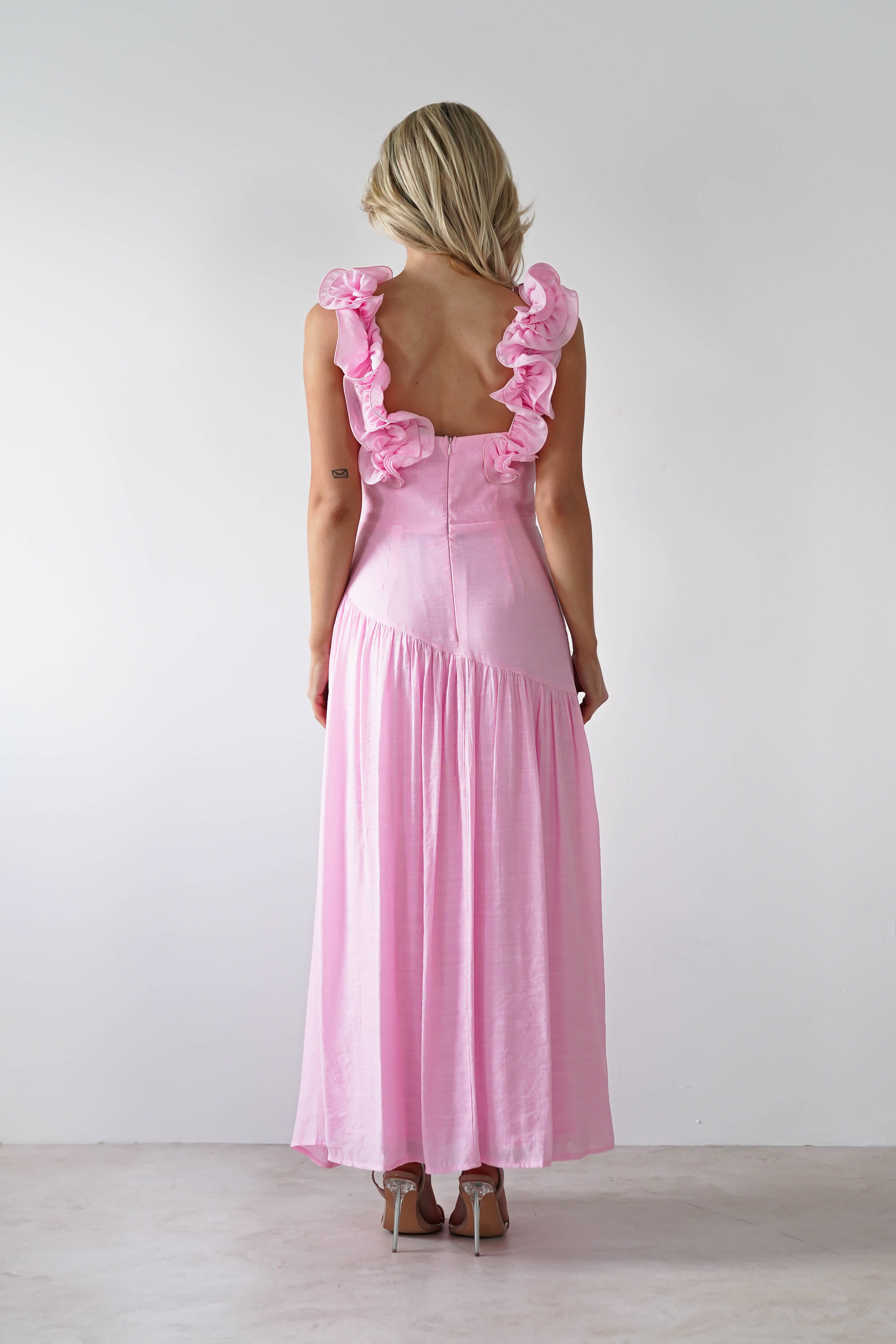 A person with blonde hair is elegantly attired in a pink midaxi dress featuring ruffled sleeves and an open back. They stand on a light-colored floor, facing away from the camera, against a plain white backdrop—ideal attire for stylish wedding guest dresses.