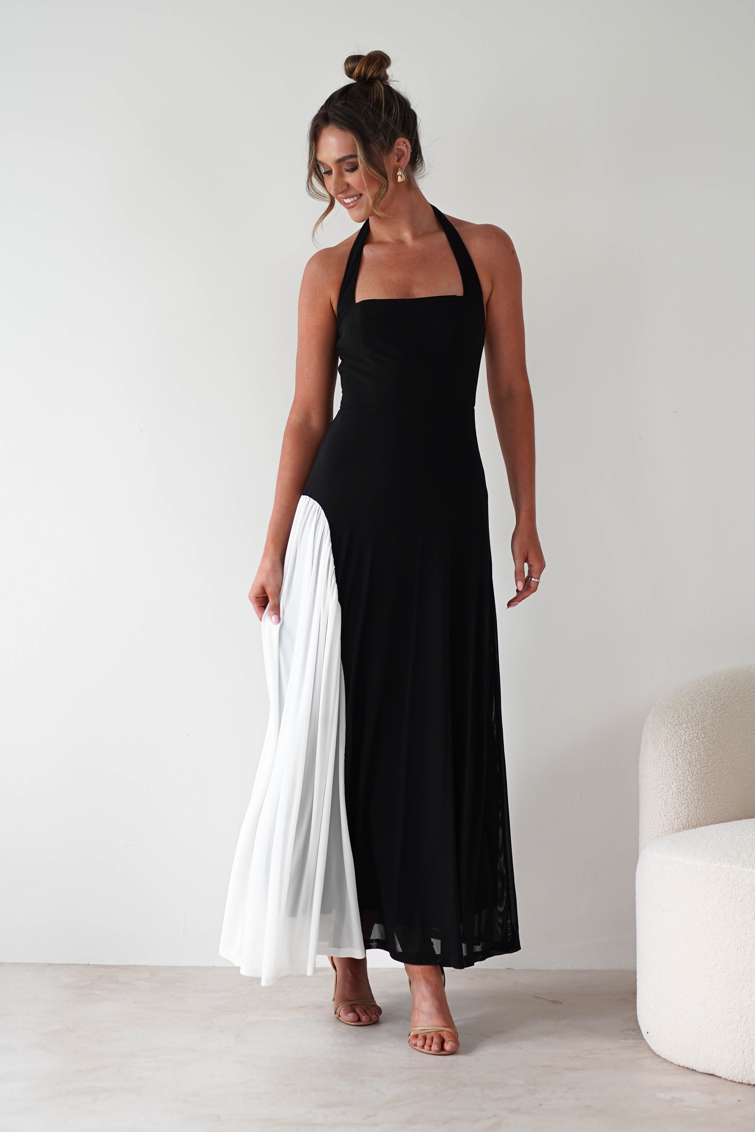 A woman in a sleeveless black halter-neck maxi dress with a white pleated panel stands, holding the skirt. With her hair up in a bun, she smiles and poses against a plain white background, showing off the comfortable fit.