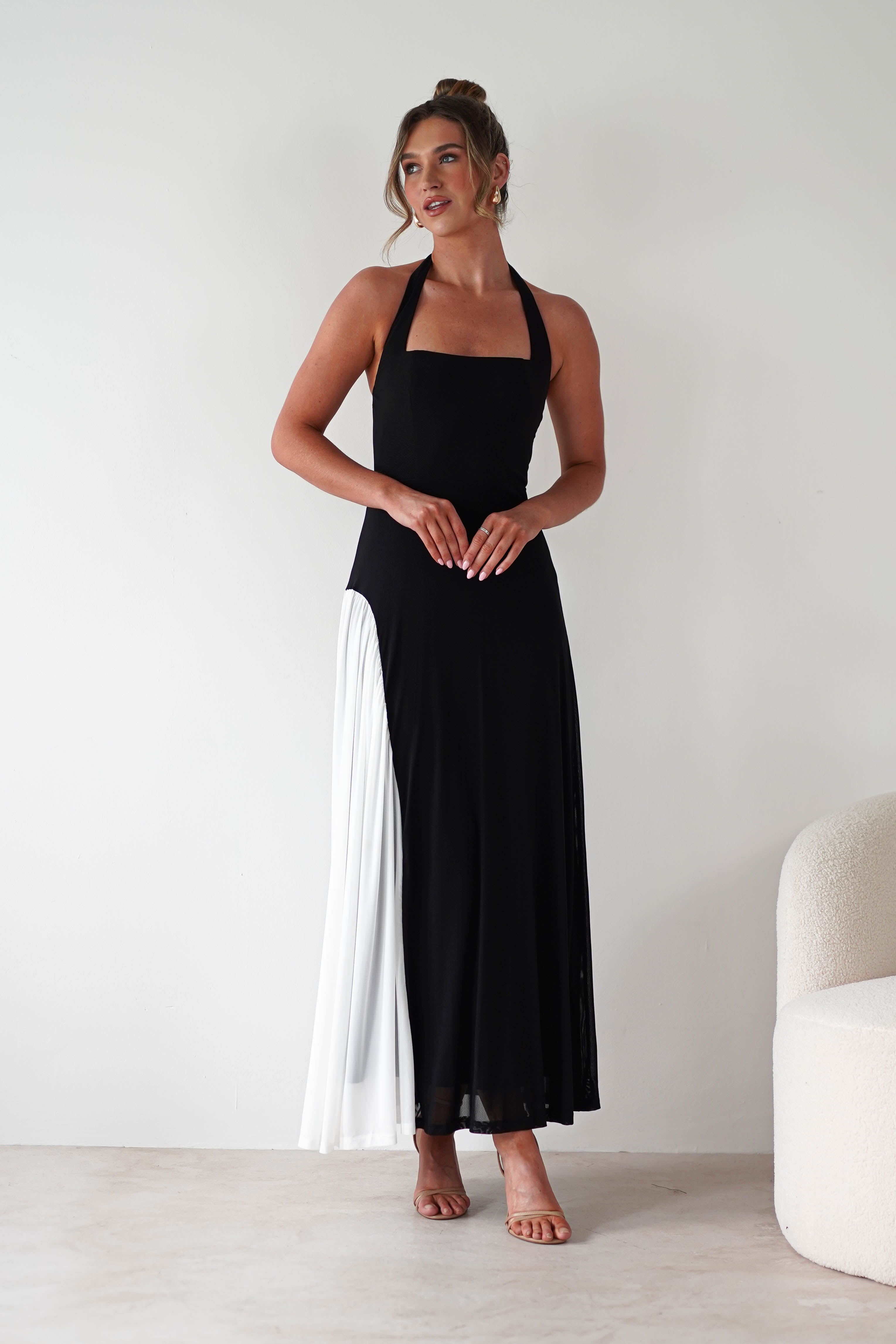 A woman stands indoors in a black halter-neck maxi dress with a square neckline and a white pleated section on one side, crafted from stretchy fabric for a comfortable fit. She pairs it with nude heels, poses with hands clasped, and gazes slightly to the side.