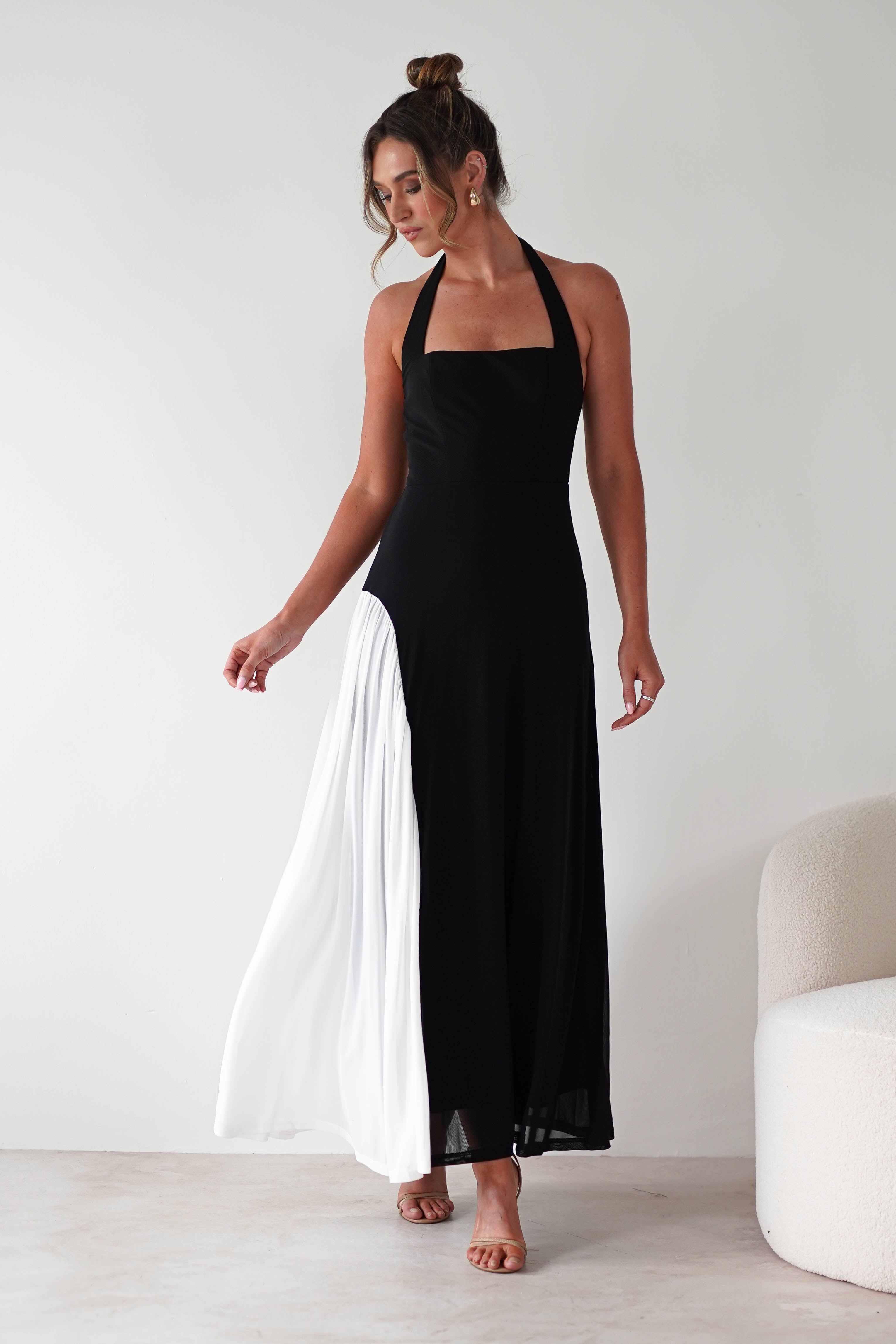 A woman stands indoors wearing a sleeveless black halter maxi dress with a white pleated section on one side. Her hair is in a loose bun, and she poses gracefully near a cream-colored chair against a plain white wall.