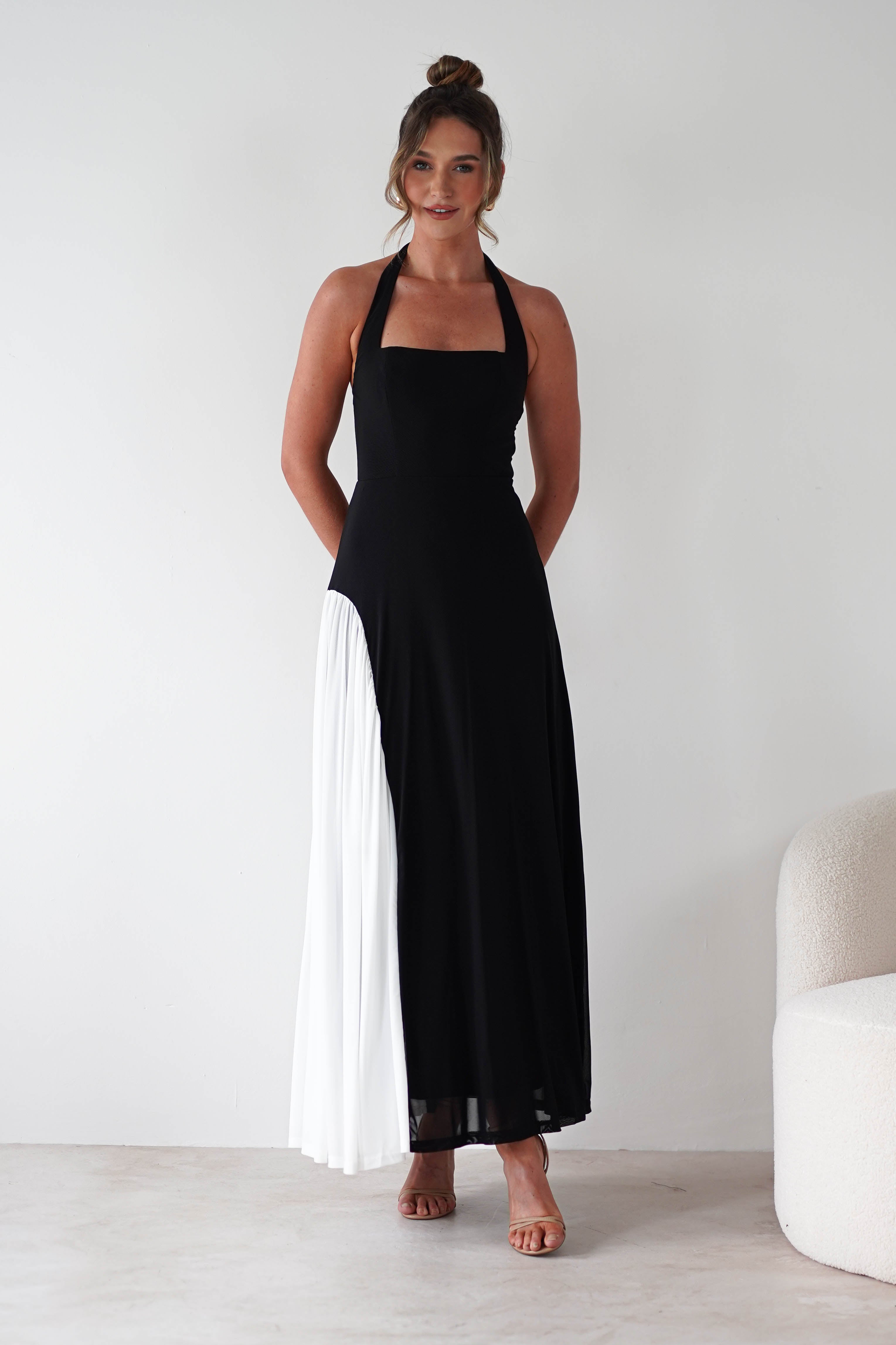 A woman wearing a sleeveless, floor-length black maxi dress with a halter neckline, a white pleated panel on one side, and a comfortable fit stands smiling in a minimalist room with a white wall and a light-colored chair.