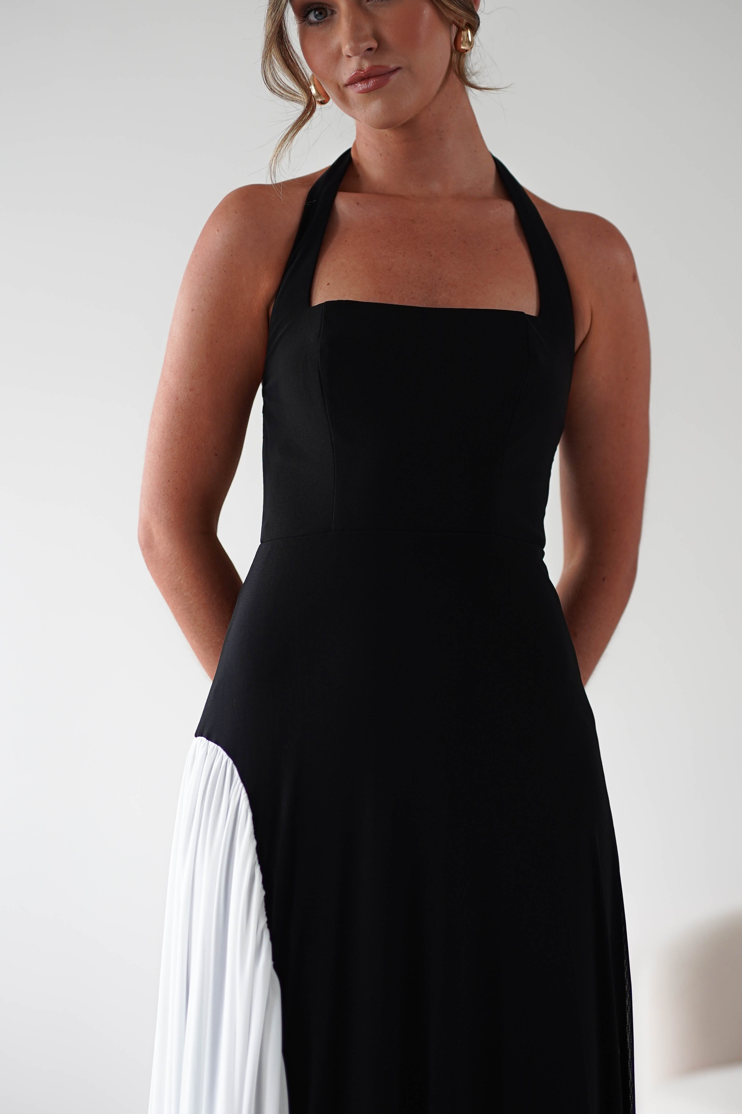A woman wears a sleeveless black maxi dress with a square neckline and white pleated detail on one side, made from stretchy fabric for a comfortable fit, standing against a plain light background with her hands behind her back.