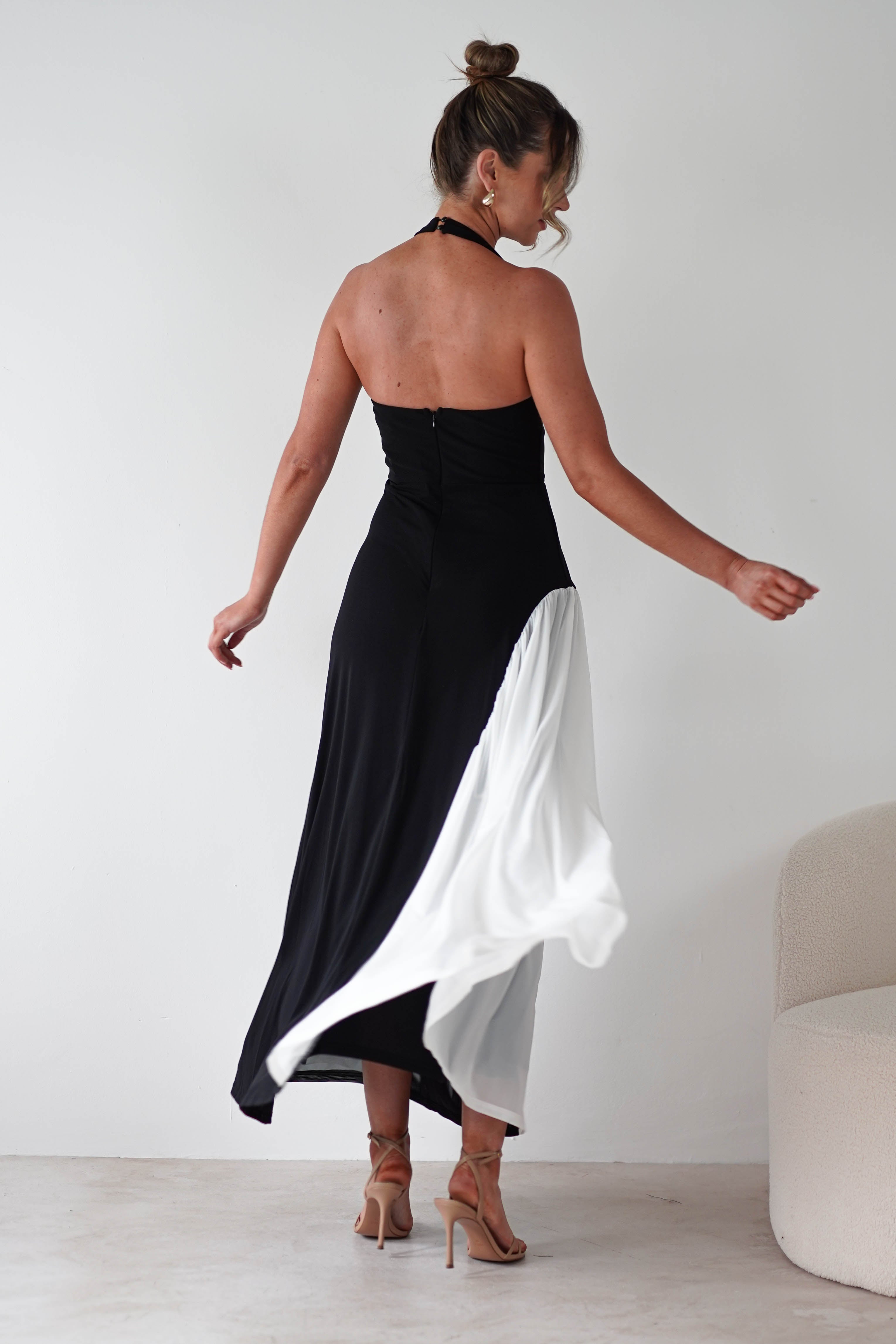 A woman twirls in a strapless black and white maxi dress, crafted with stretchy fabric for a comfortable fit. With her back to the camera, she raises one arm slightly beside a light-colored chair against a plain white background.