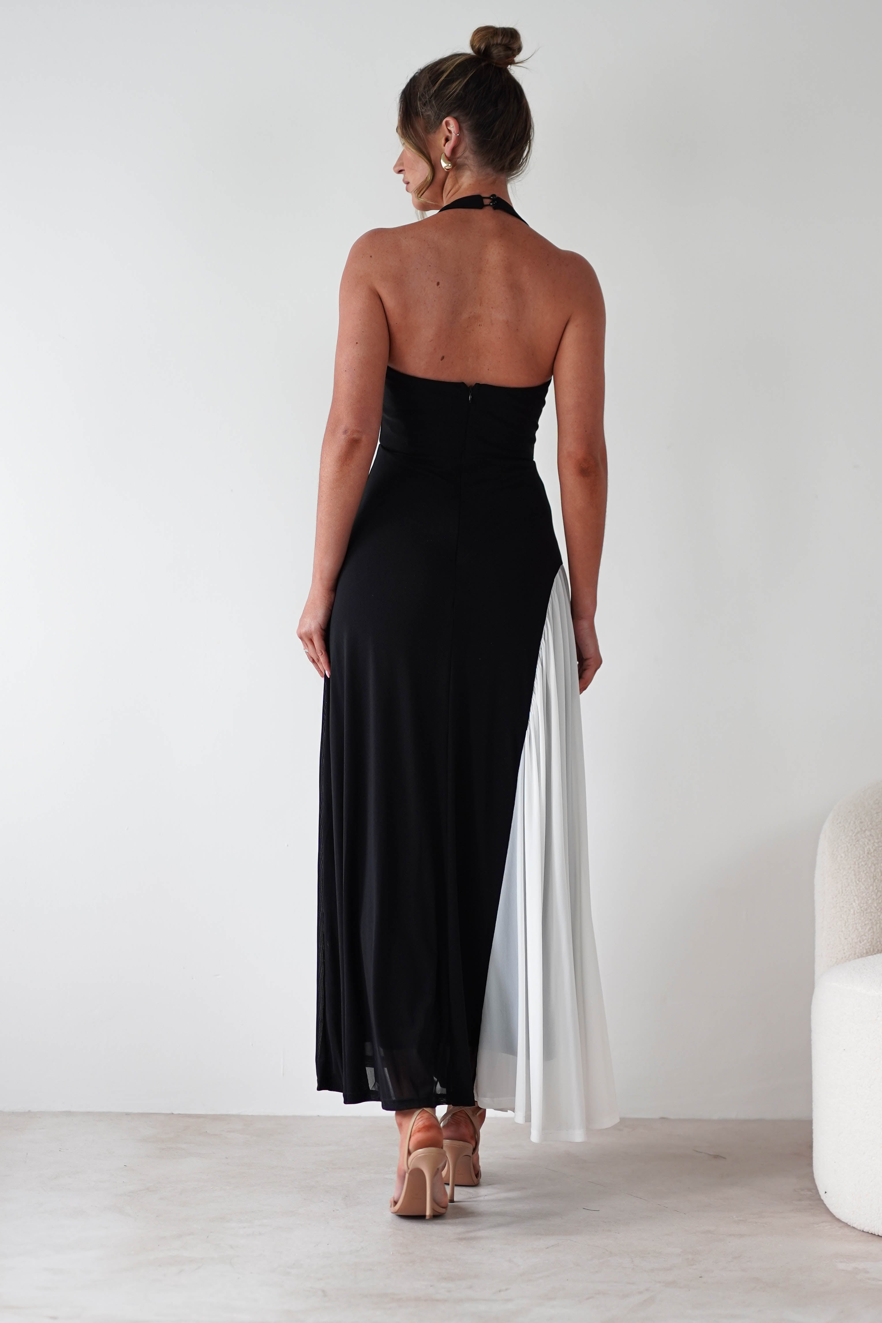 A woman stands facing away, wearing a black strapless maxi dress with a white pleated panel on one side. Made from stretchy fabric for a comfortable fit, her hair is in a bun and she wears nude heels against a plain white background.