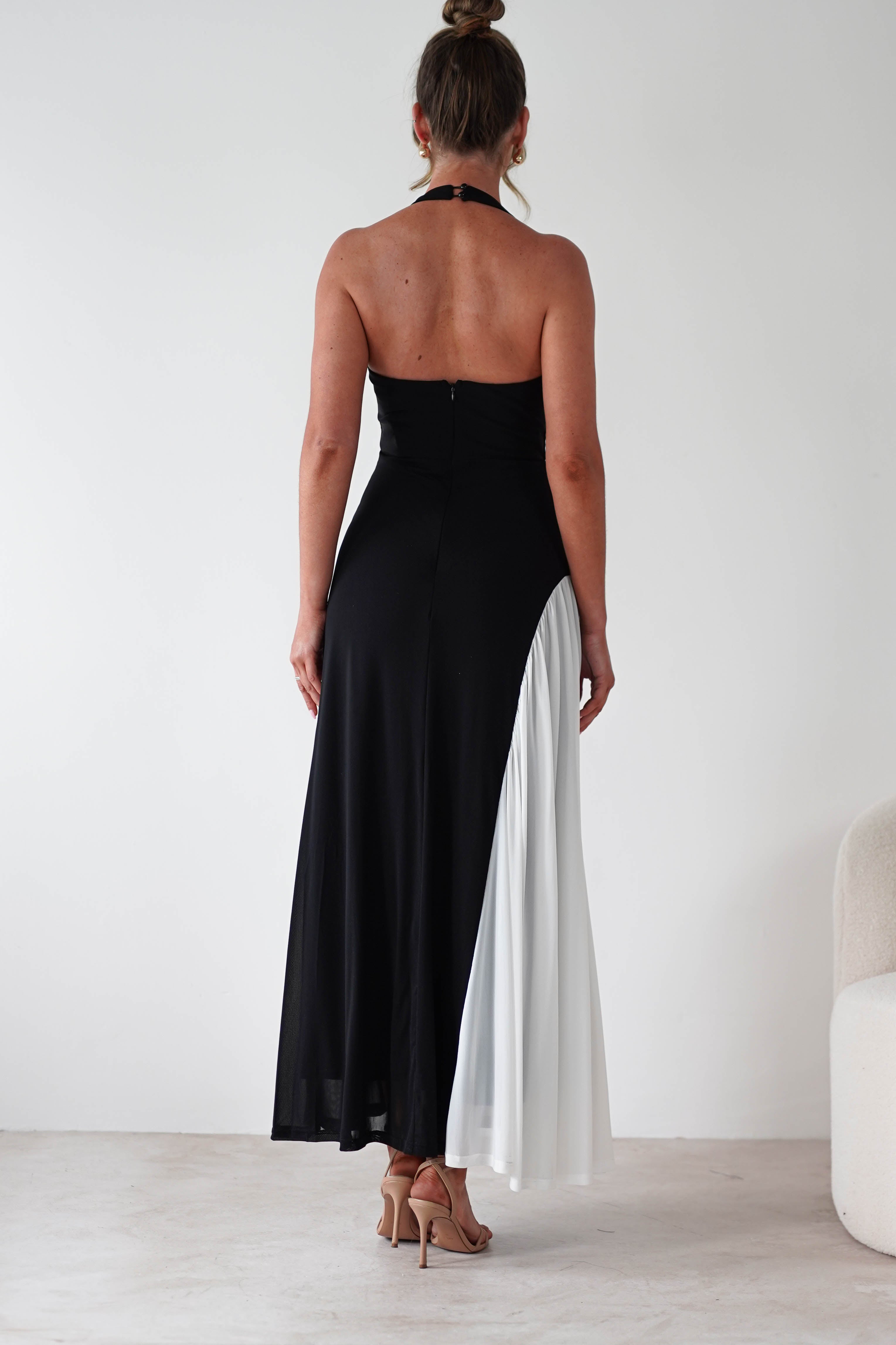 A woman stands facing away, wearing a black halter-neck maxi dress with a white pleated panel on one side and nude high heels. The stretchy fabric offers a comfortable fit, and her hair is in a bun as she stands in a minimal, light-colored room.