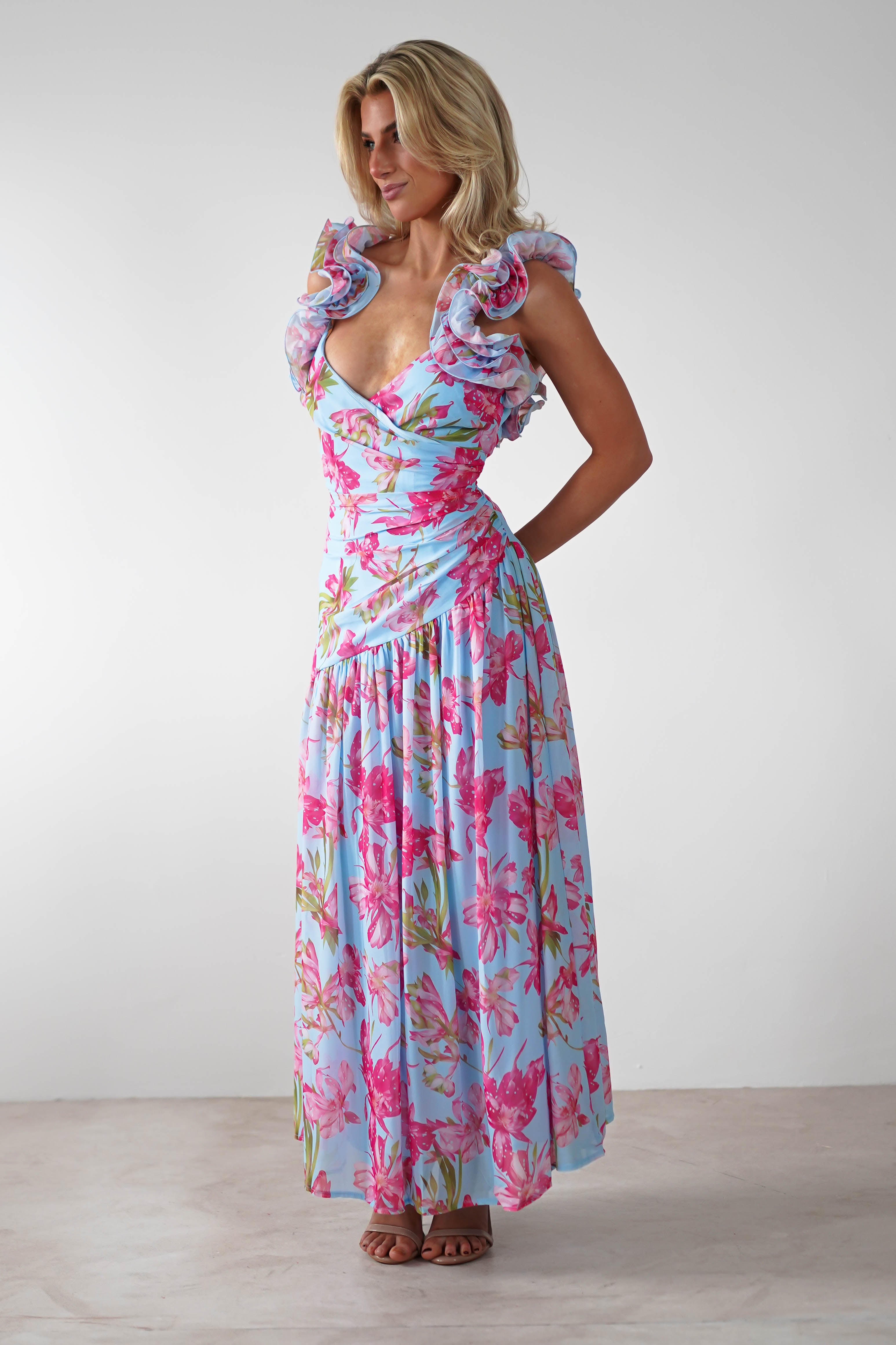 A woman stands indoors wearing a vibrant floral print ruffle shoulder maxi dress in light blue and pink. She has blonde hair, is smiling, and has her hands behind her back—the perfect look for a wedding guest.