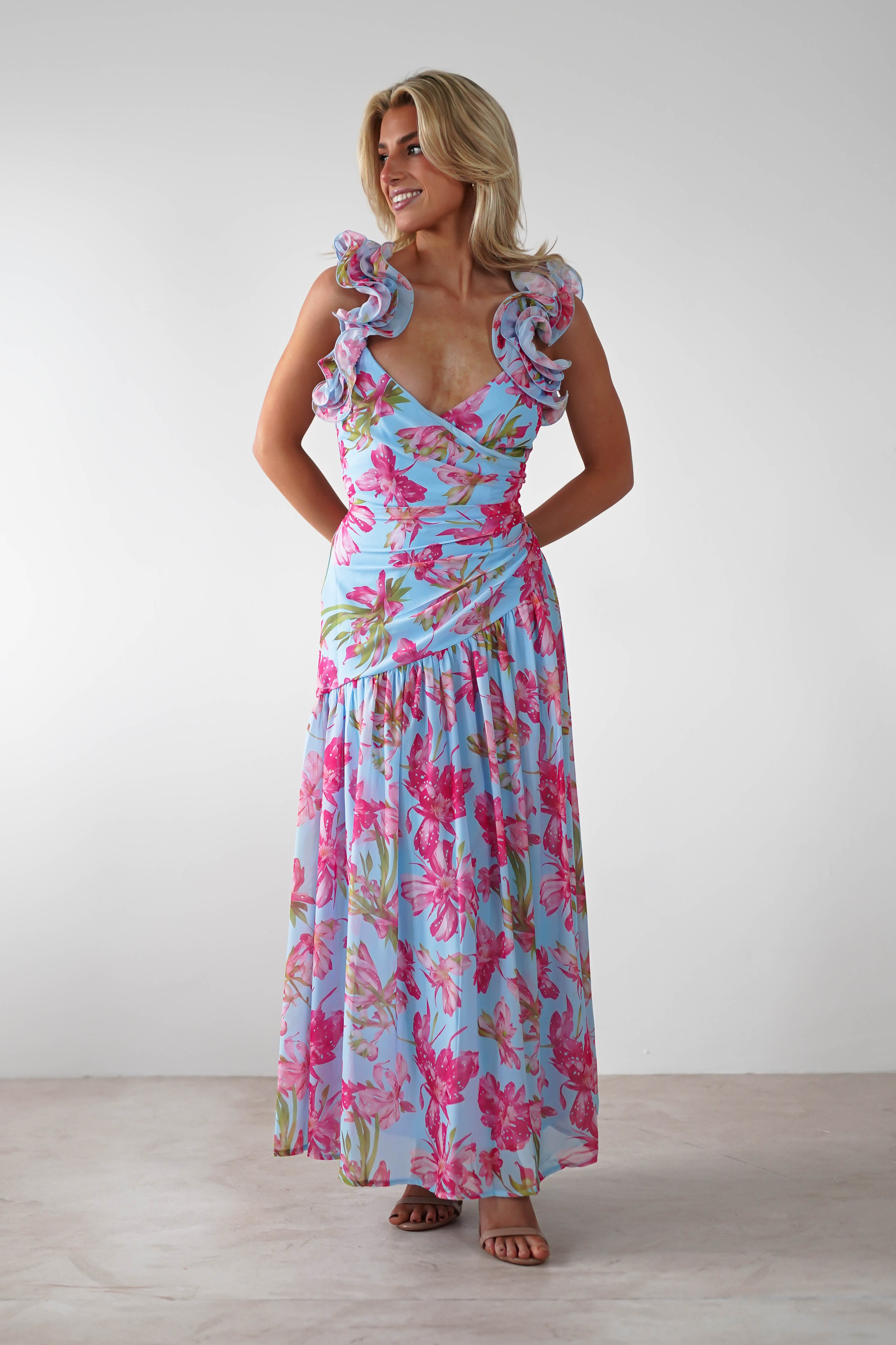 A woman stands smiling in a vibrant floral print ruffle shoulder maxi dress, perfect for a wedding guest. The light blue, floor-length dress features pink and green patterns and ruffled sleeves. She poses with blonde hair and hands behind her back.