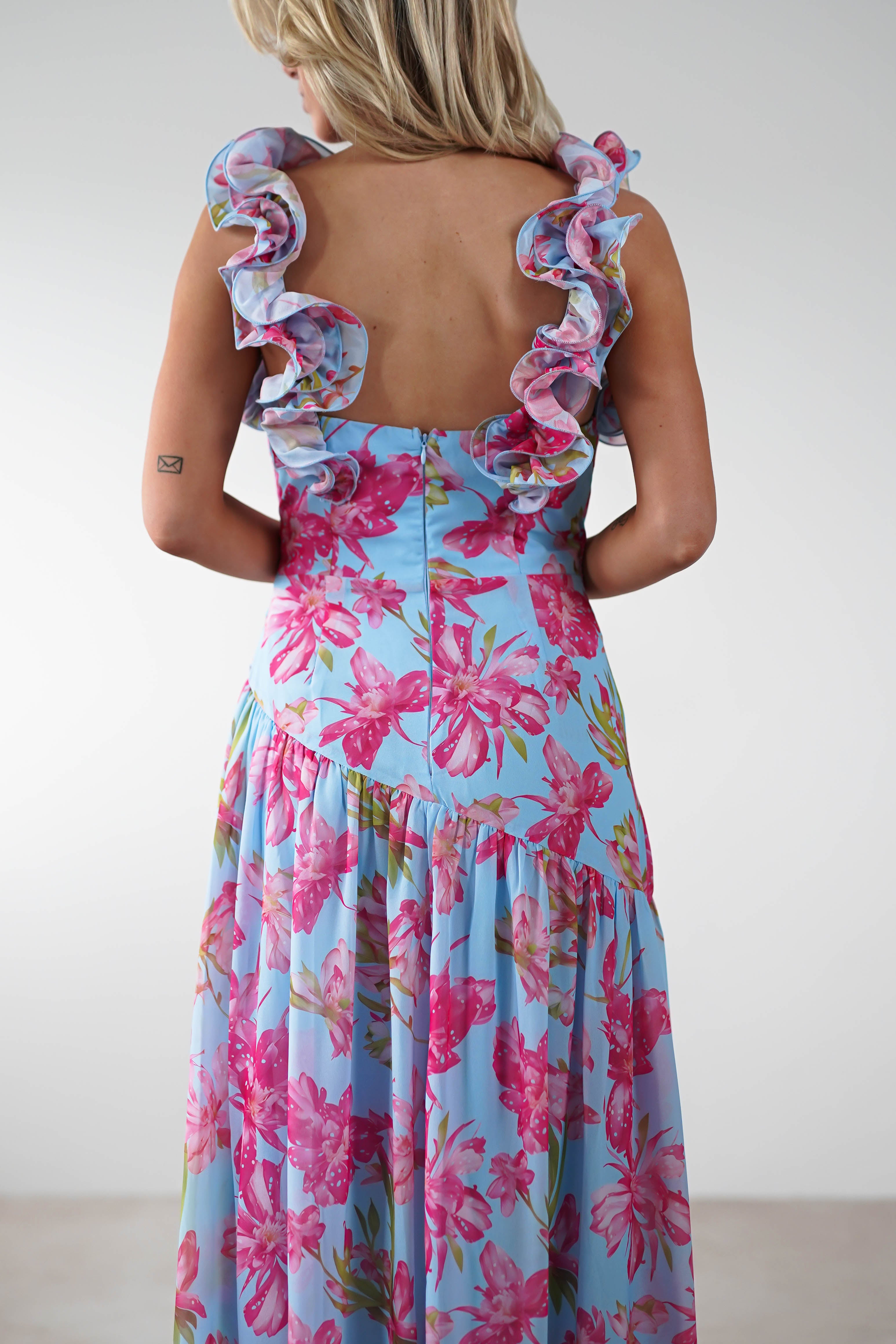 A woman with blonde hair faces away, wearing a vibrant floral print ruffle shoulder maxi dress with a fitted bodice and flowing skirt—perfect for a stylish wedding guest. A small tattoo is visible on her right arm.
