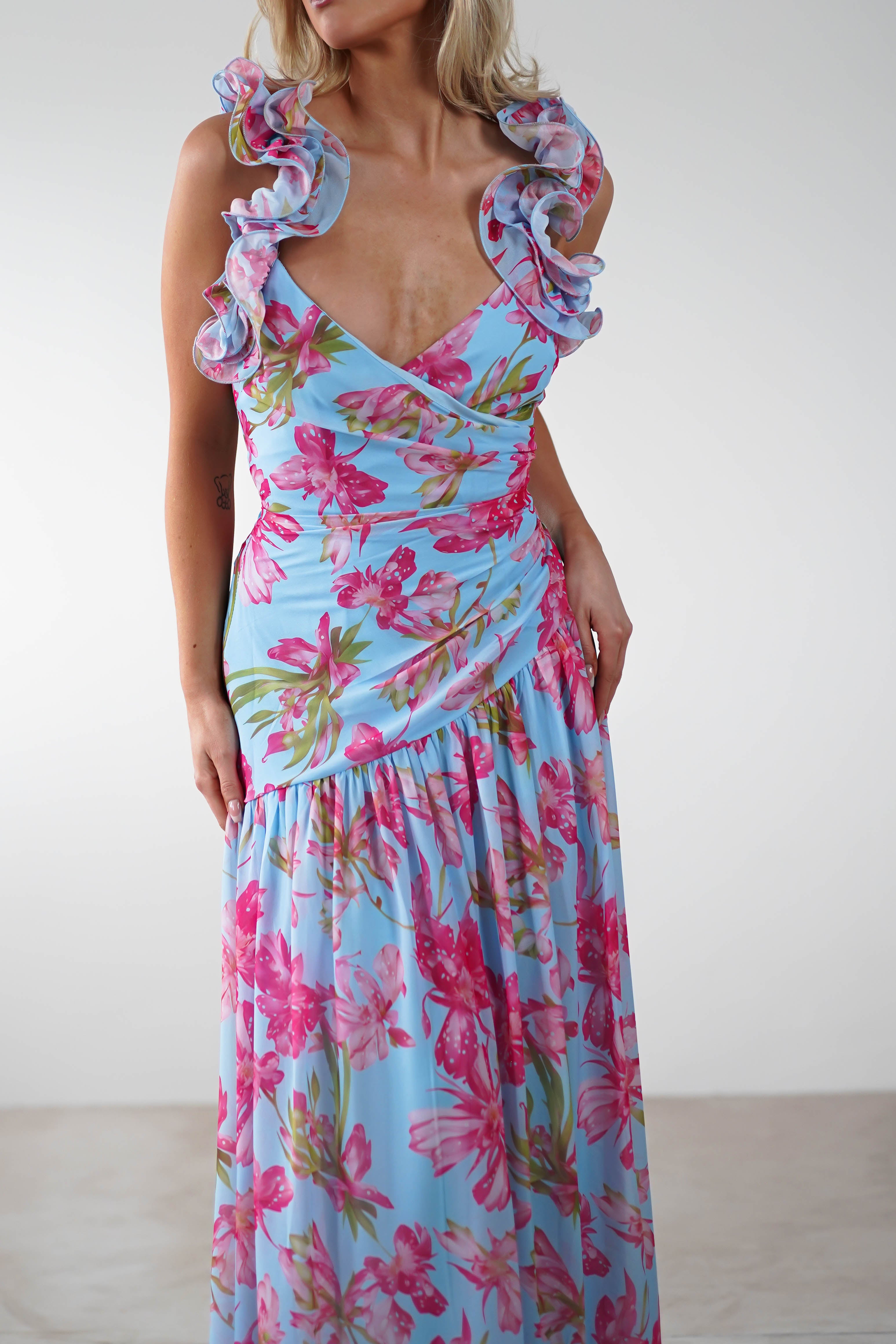 A woman wears a light blue ruffle shoulder maxi with vibrant floral print, featuring a fitted bodice and v-neckline. The background is plain and light-colored—perfect as a wedding guest look.