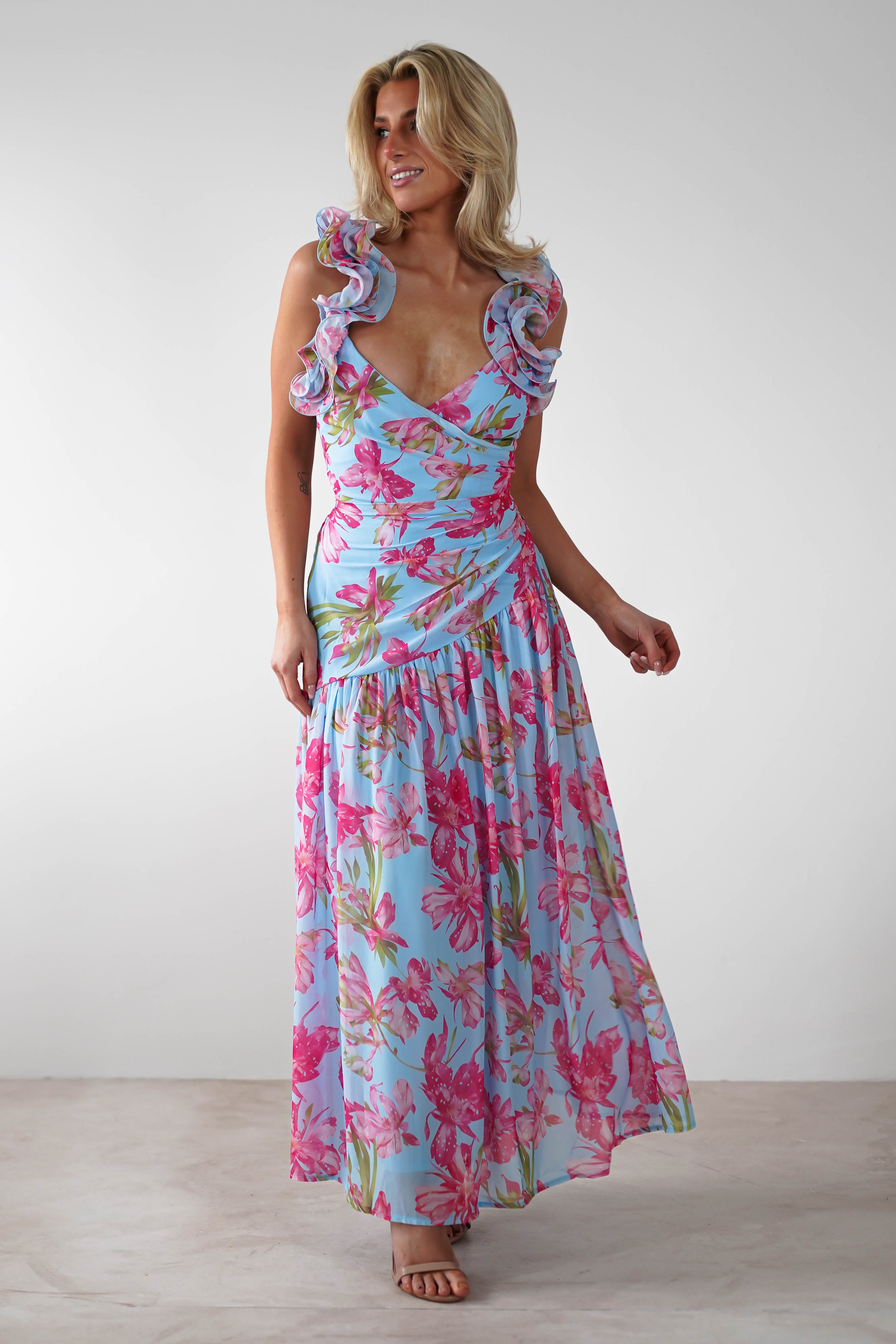A woman wearing a vibrant floral print ruffle shoulder maxi dress in light blue with pink and green accents stands in a neutral setting, smiling and looking to the side—perfect as a wedding guest outfit.