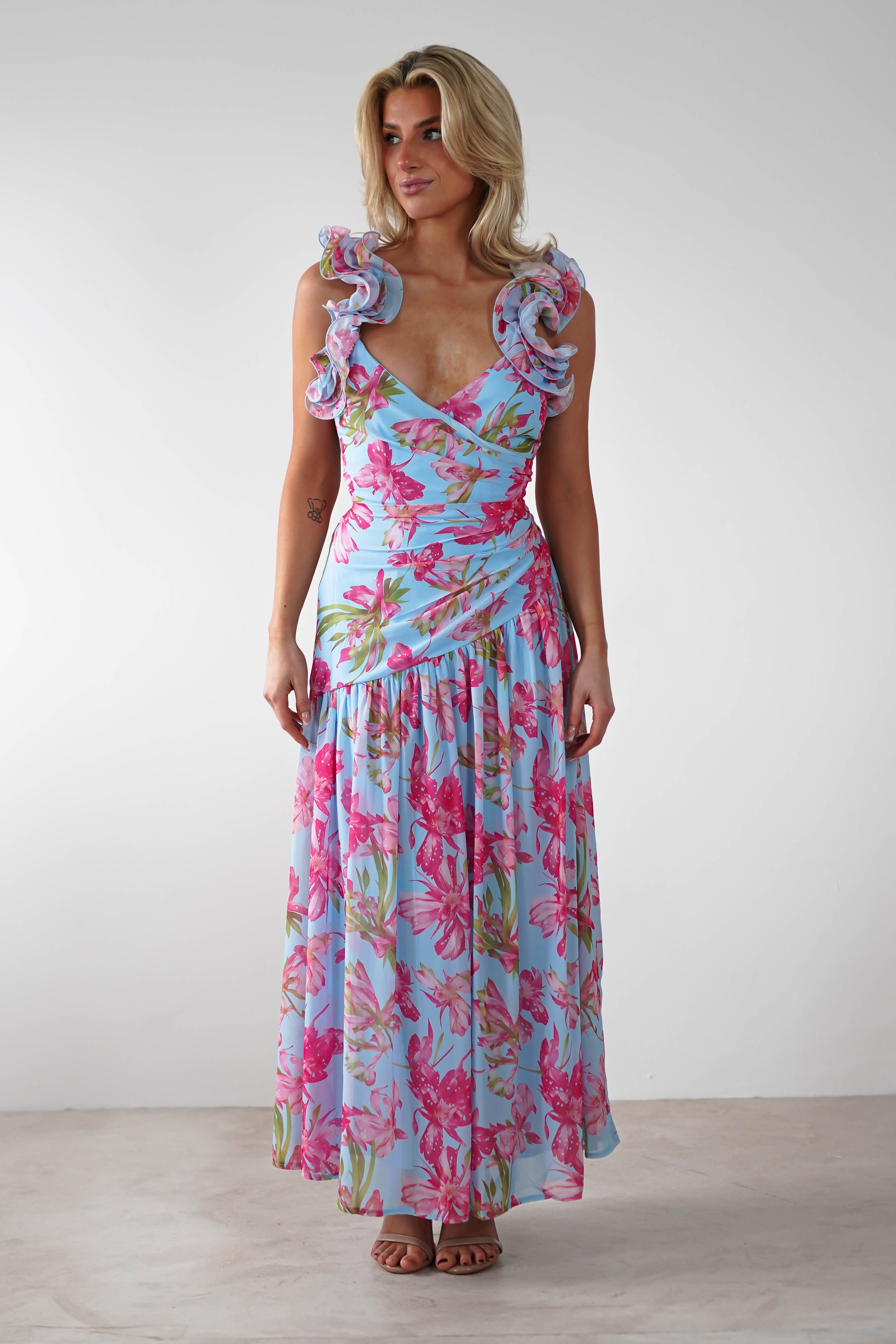 A woman wearing a vibrant floral print ruffle shoulder maxi dress in blue with pink and green accents stands against a plain light background, perfect for a wedding guest look.