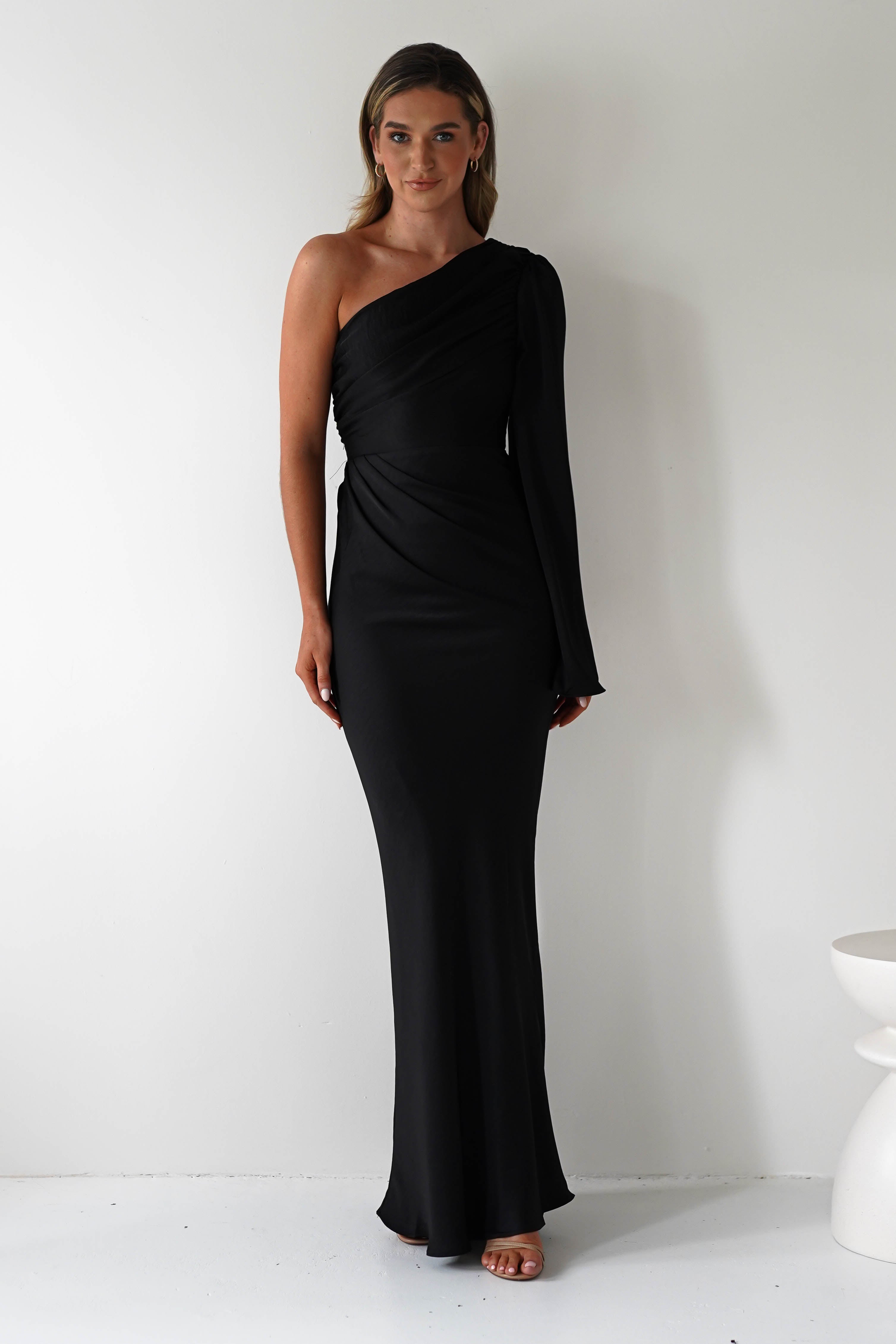 A woman stands against a plain white background wearing an elegant ensemble featuring a sleek, one-shoulder black gown. The asymmetrical neckline complements the long, flowing silhouette, as her long hair cascades gracefully. She stands with one arm by her side, smiling softly.