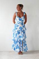 Woman wearing a white maxi dress with blue floral print, ruffled shoulder straps, and tiered skirt, standing against a plain white background.