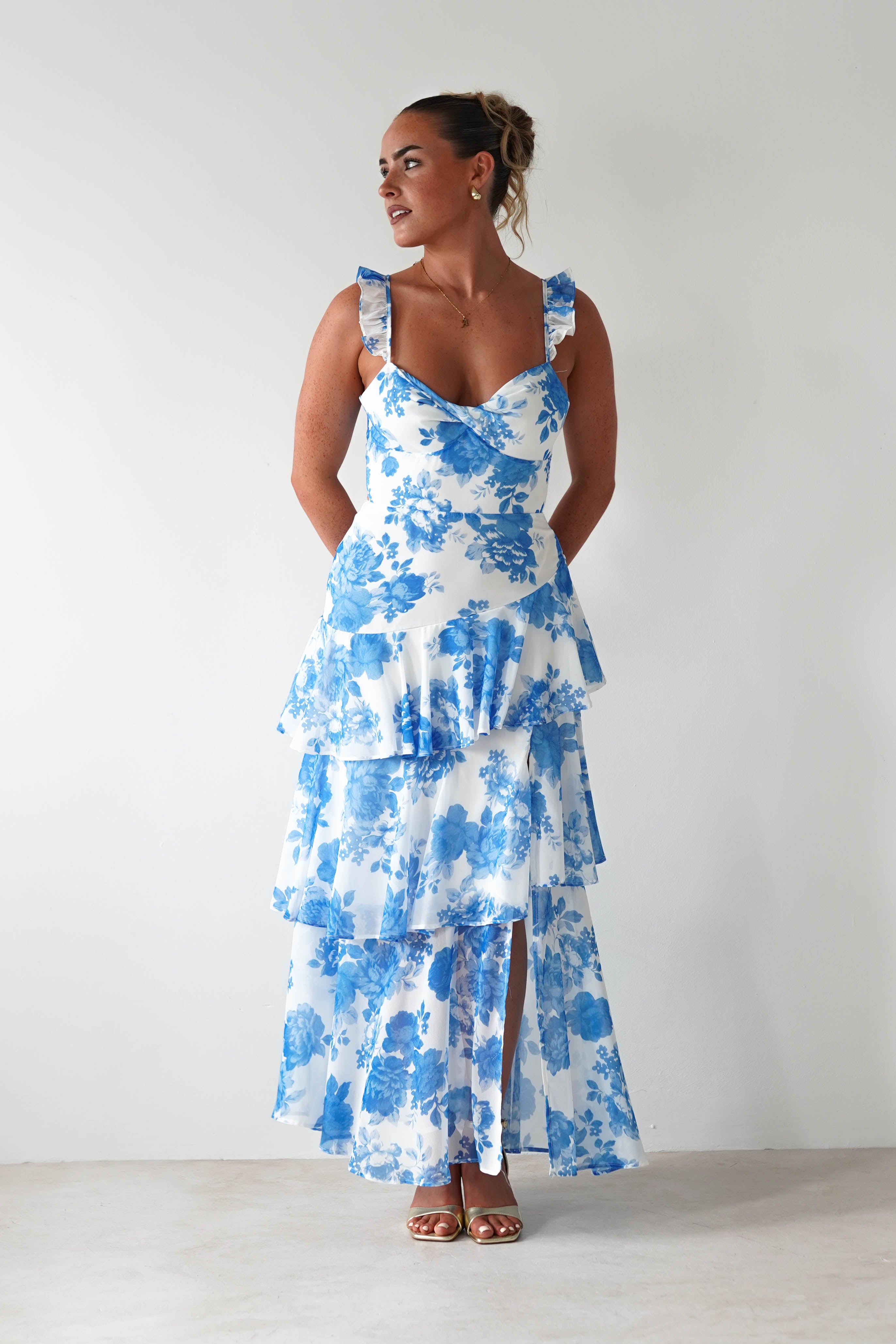 Woman wearing a white maxi dress with blue floral print, ruffled shoulder straps, and tiered skirt, standing against a plain white background.