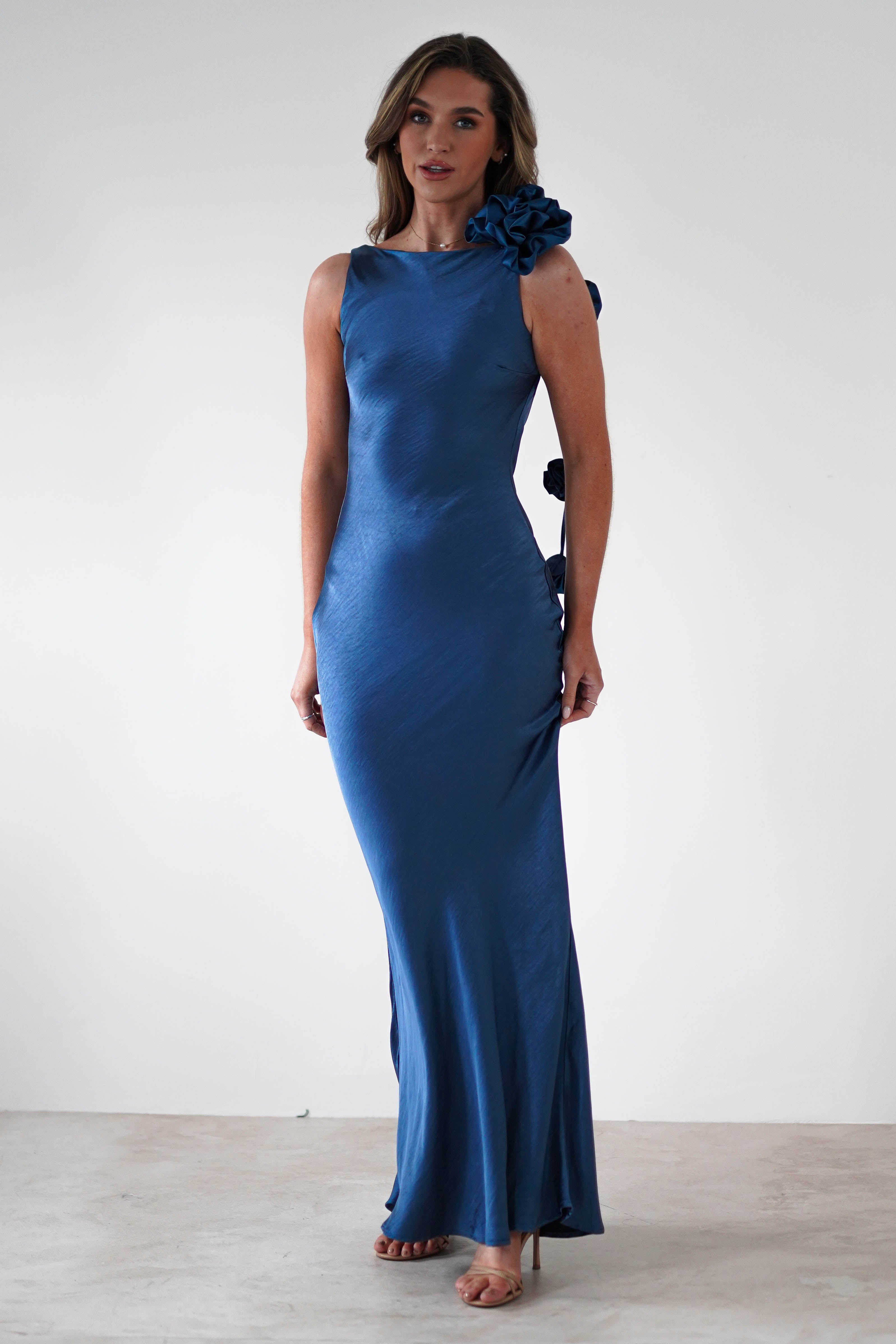 A person wearing a long, elegant blue satin dress with a floral embellishment on the shoulder and a draped flower trim stands against a plain white background, looking at the camera. The dress features a bateau neckline, slim fit, and slight train.