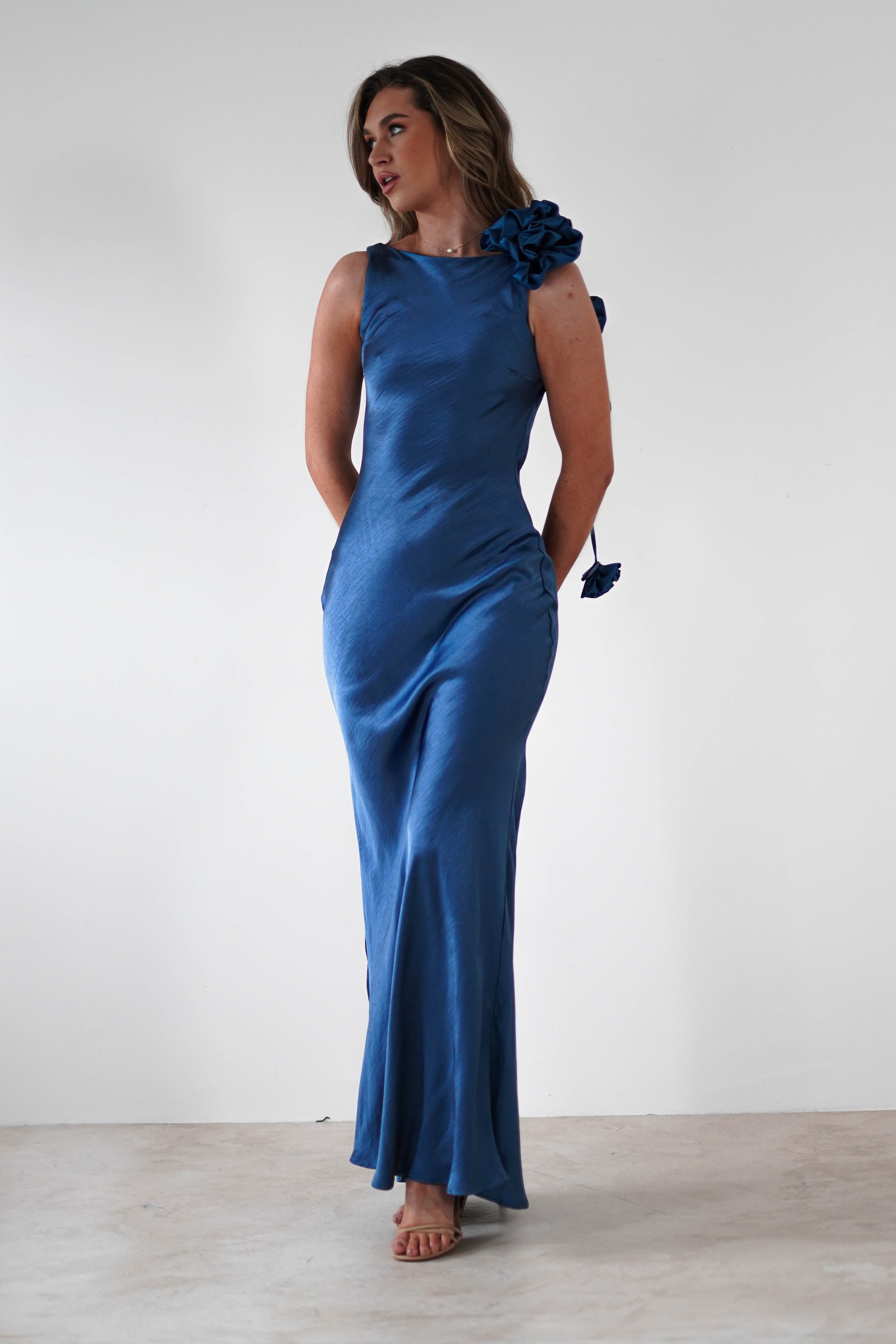 A woman stands sideways, hands behind her back, in a long, shiny blue dress featuring a draped flower trim on one shoulder. The gown boasts a sophisticated bateau neckline. Her long, wavy hair complements her elegant profile against the plain white background.