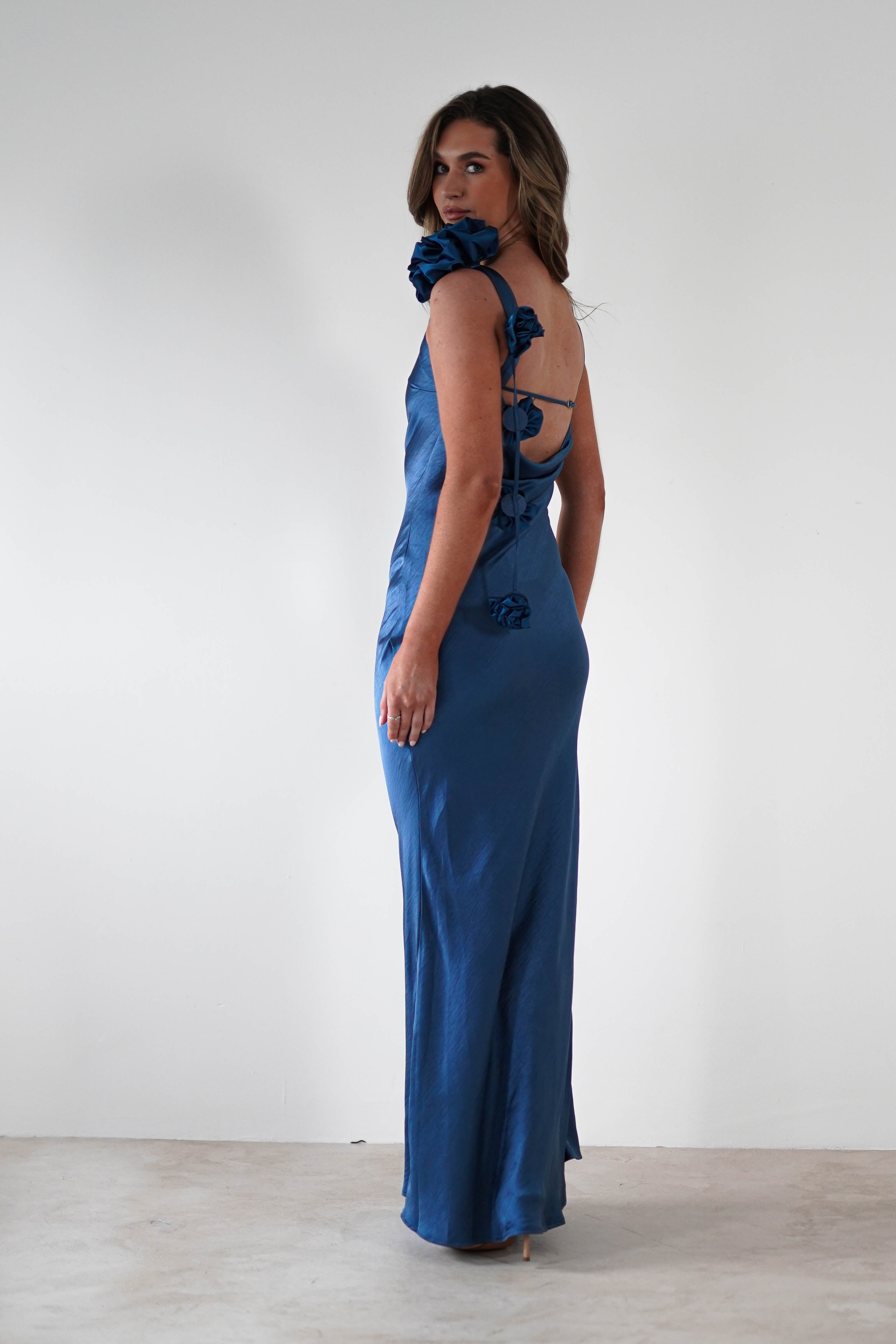 A person dons an elegant, sleeveless blue satin gown, showcasing floral accents and a bateau neckline on the shoulder straps. The dress also features a graceful cowl back, with delicate straps against a plain white backdrop.