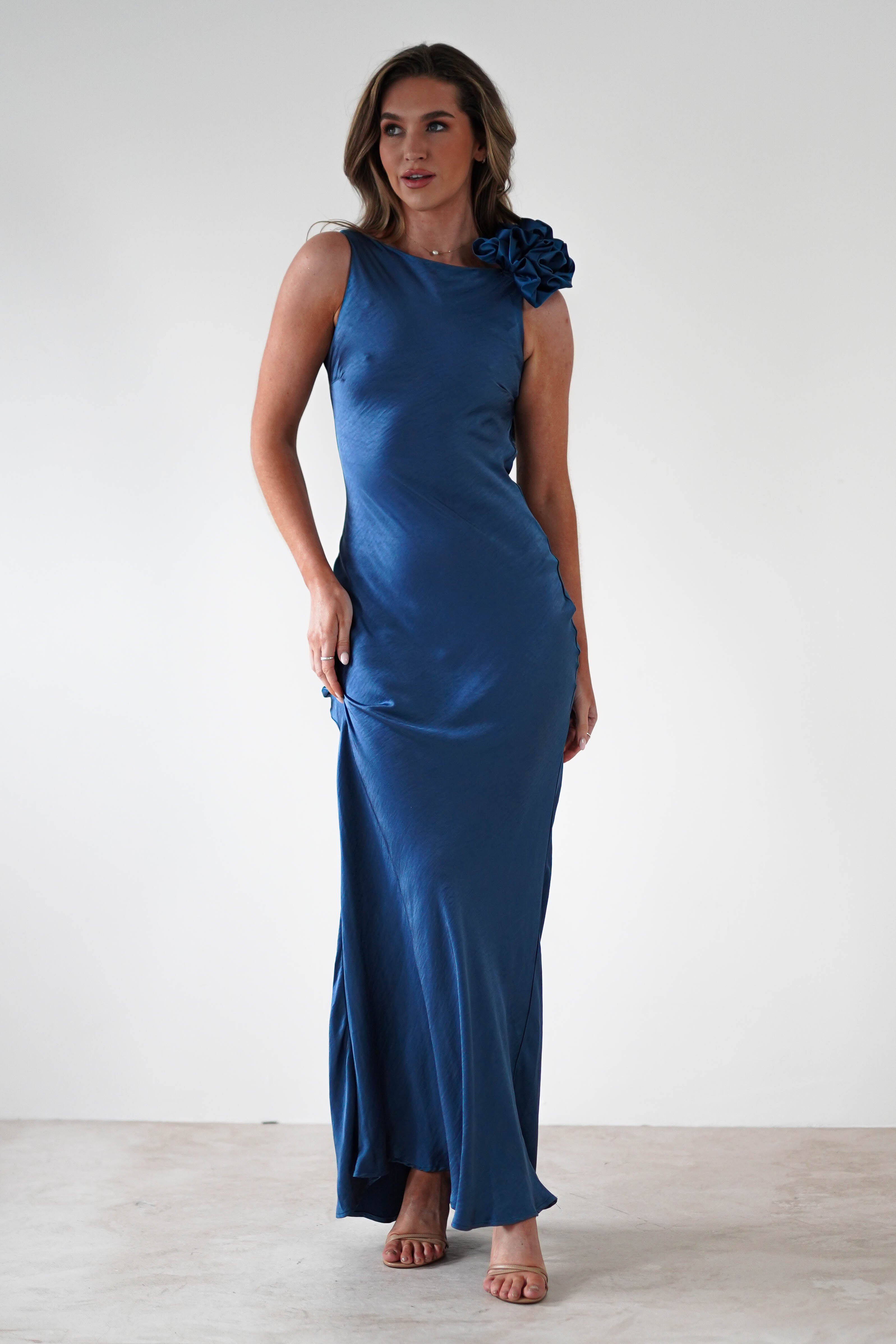 A person elegantly wears a long, sleeveless blue satin dress featuring a draped flower trim on one shoulder. They stand confidently on a light-colored floor against a plain white background, with one hand resting on their hip and the other at their side.
