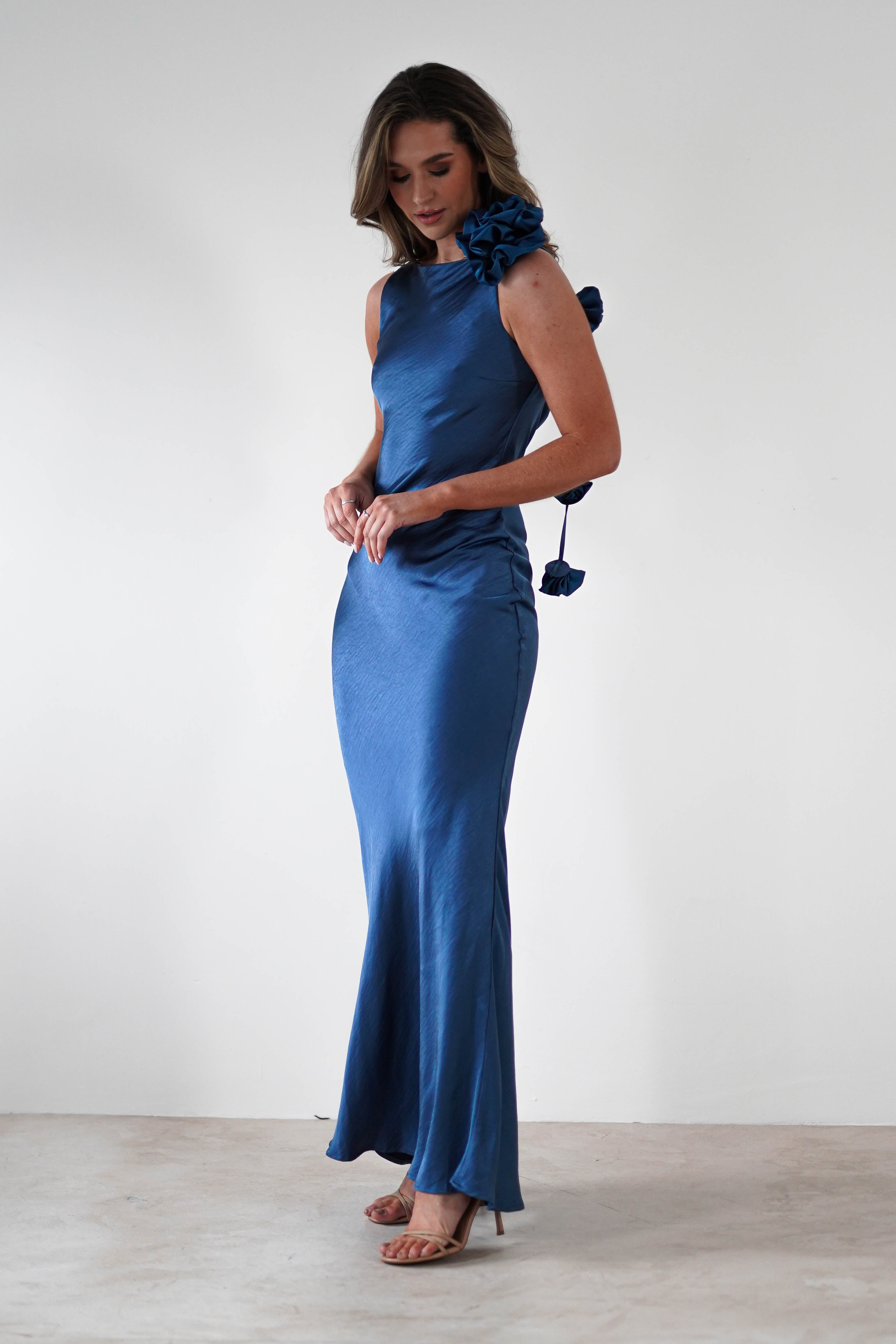 A woman stands in a minimalistic setting, wearing a sleeveless satin blue gown with a cowl back and draped flower trim on the shoulder. She gazes downward, softly touching the gown with her hands. Her shoes are beige.