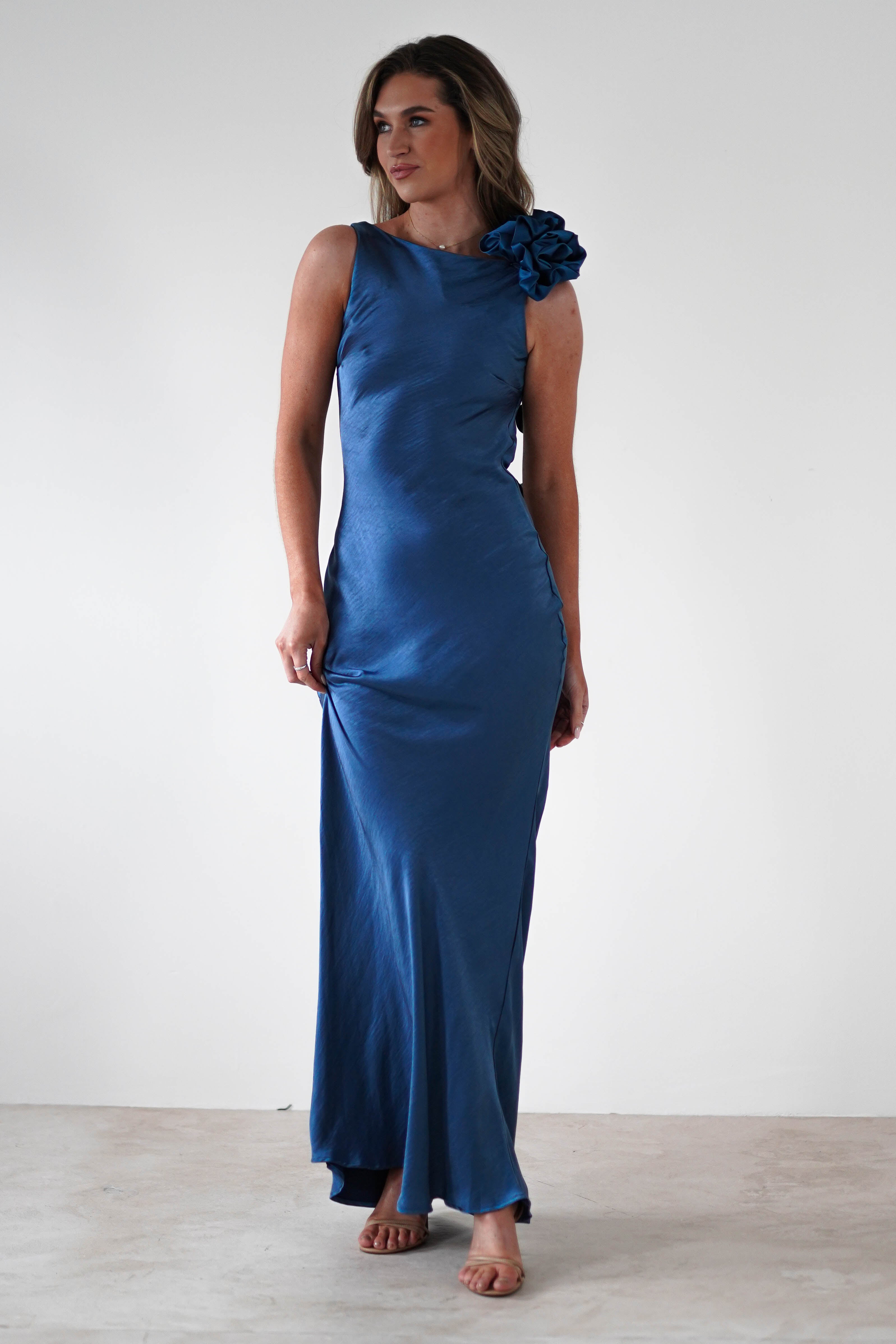 A woman wearing a sleek, sleeveless blue satin dress with a bateau neckline and floral detail on the shoulder stands against a plain white background. She gazes slightly to the side, her hands gently holding her dress adorned with an elegant draped flower trim.