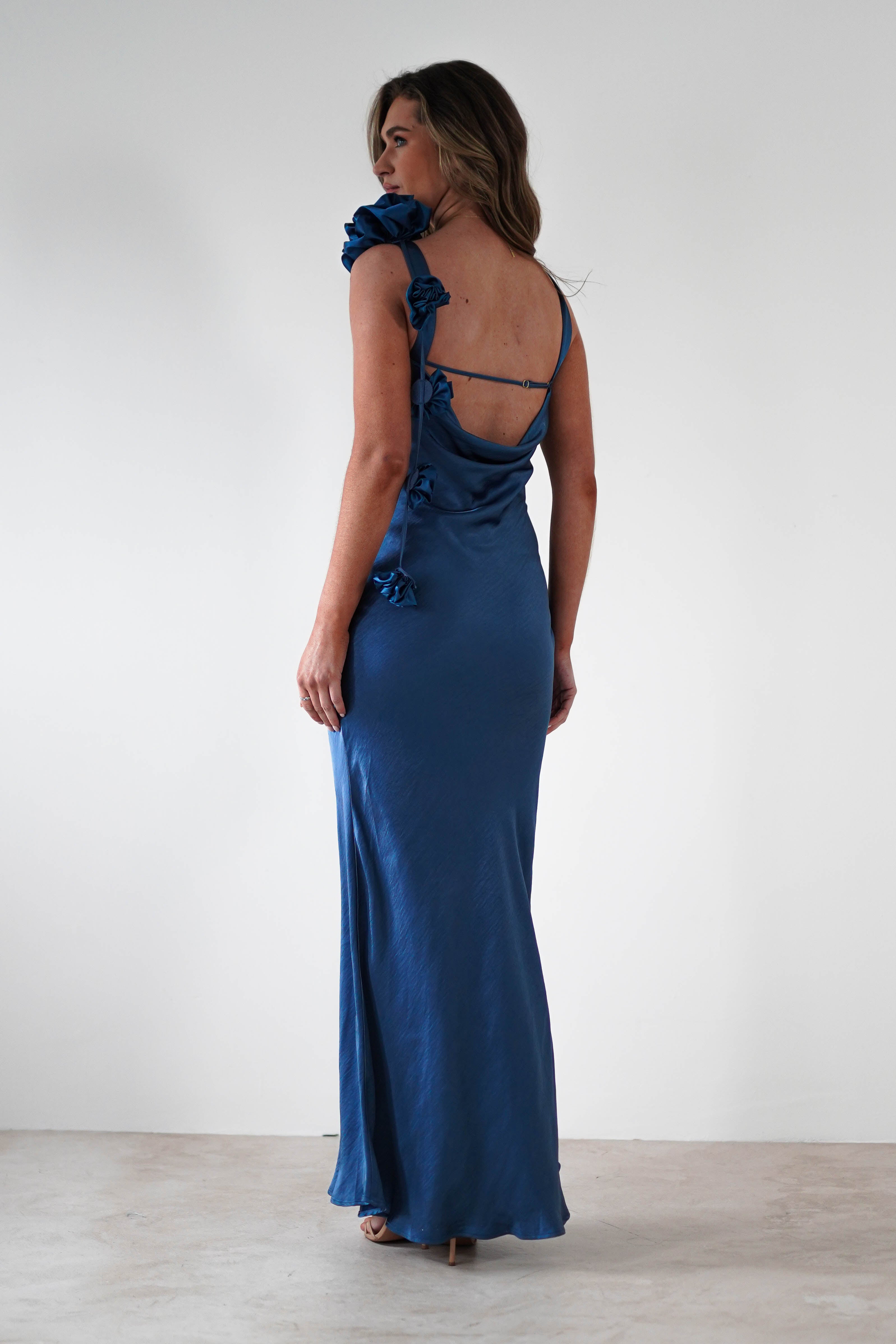 A woman in a long, elegant blue dress with a draped flower trim and a cowl back stands against a plain white background, looking over her shoulder.