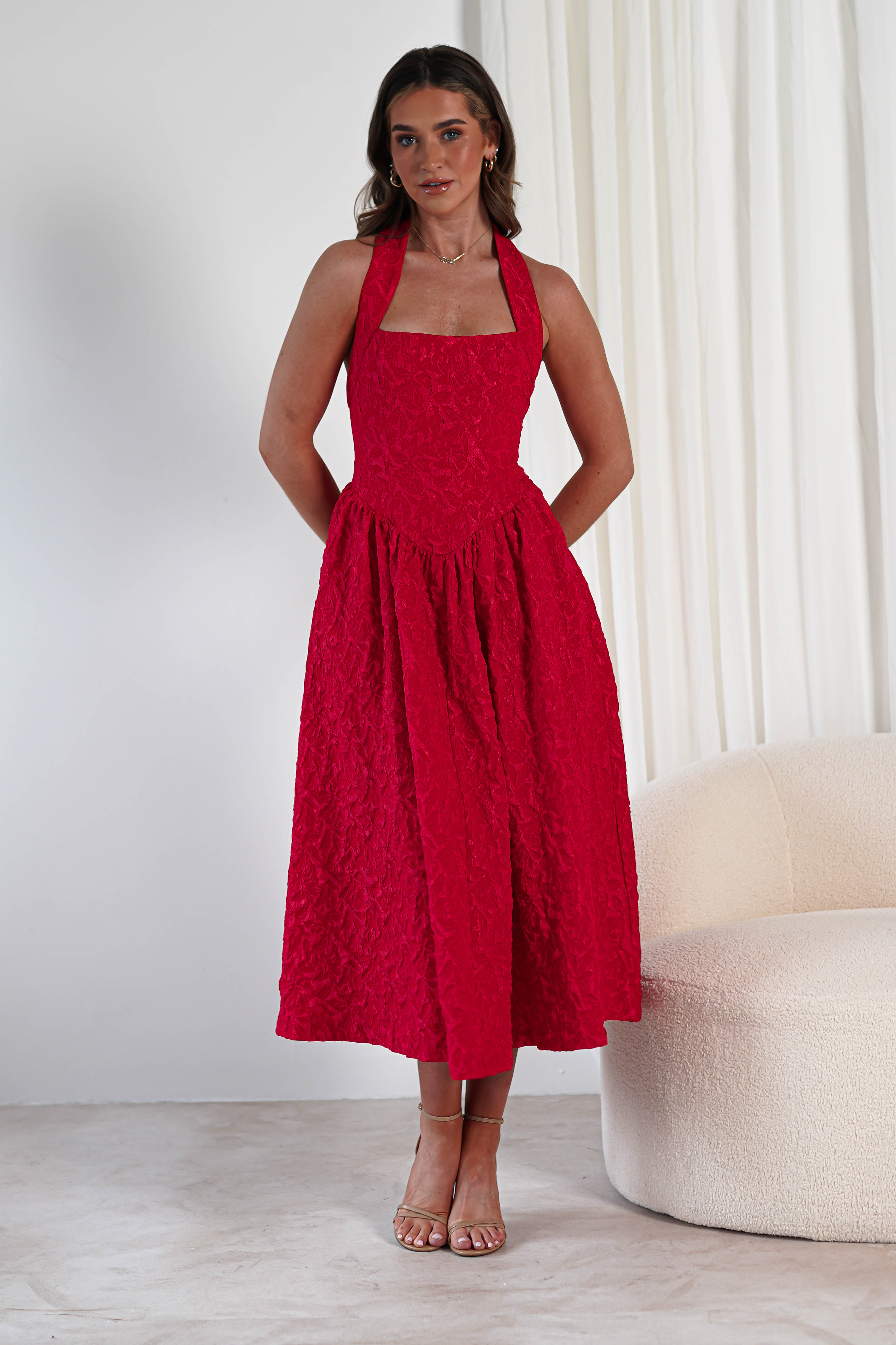A woman stands indoors wearing a sleeveless, halter-neck red midi dress crafted from textured fabric, featuring a fitted bodice, flared skirt, and practical pockets—an elegant choice for formal events. White curtains and a cream chair are in the background.