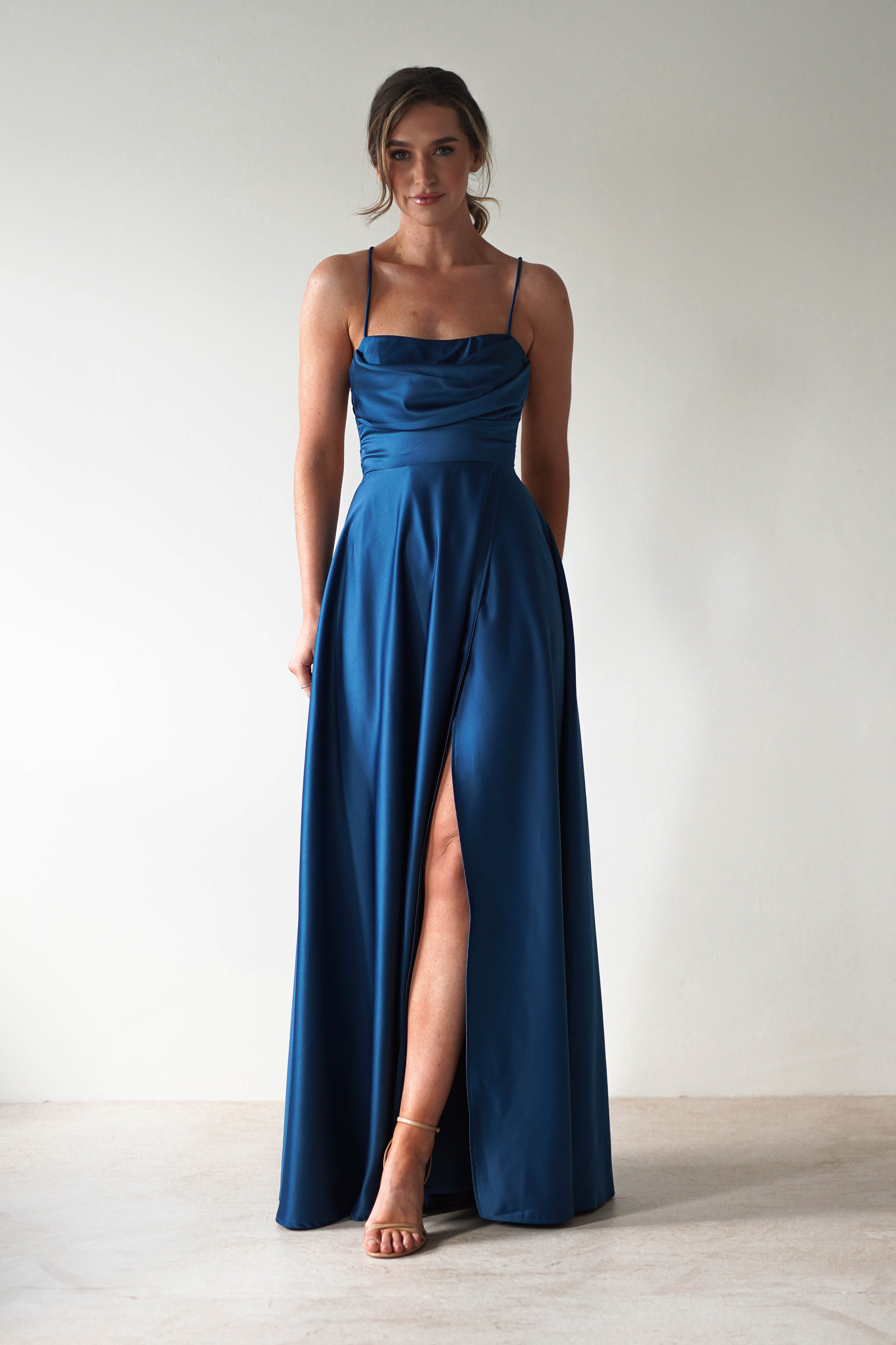 Woman in a teal satin gown with spaghetti straps and a flowing cowl neckline, standing against a plain background. The dress features a thigh-high slit that complements her elegant posture, while her hair is styled up.