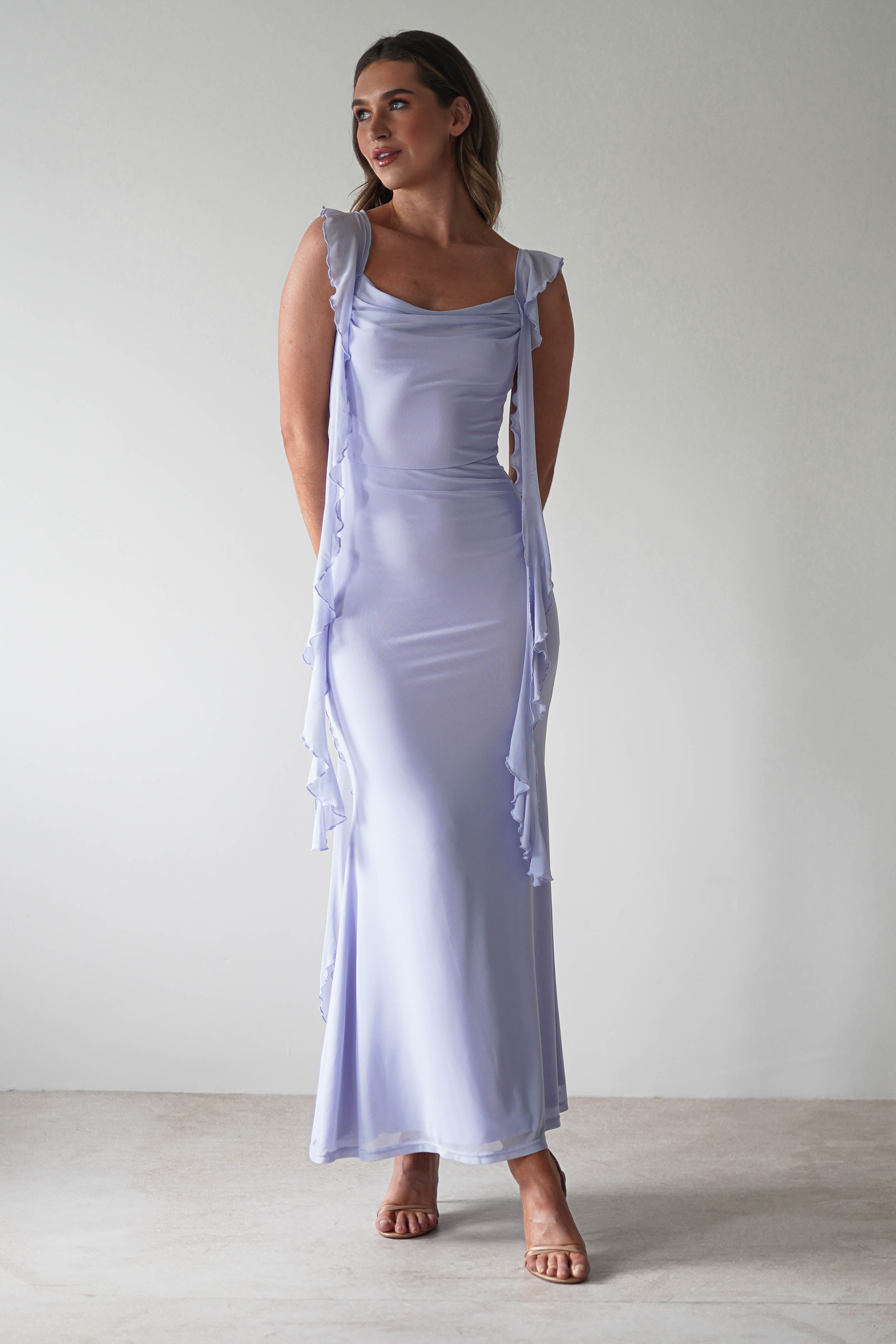 A woman stands against a plain background, wearing a light lavender chiffon dress with ruffle detail along the straps and sides. Barefoot, with long brown hair, she looks slightly to her left—perfect as a wedding guest ensemble.