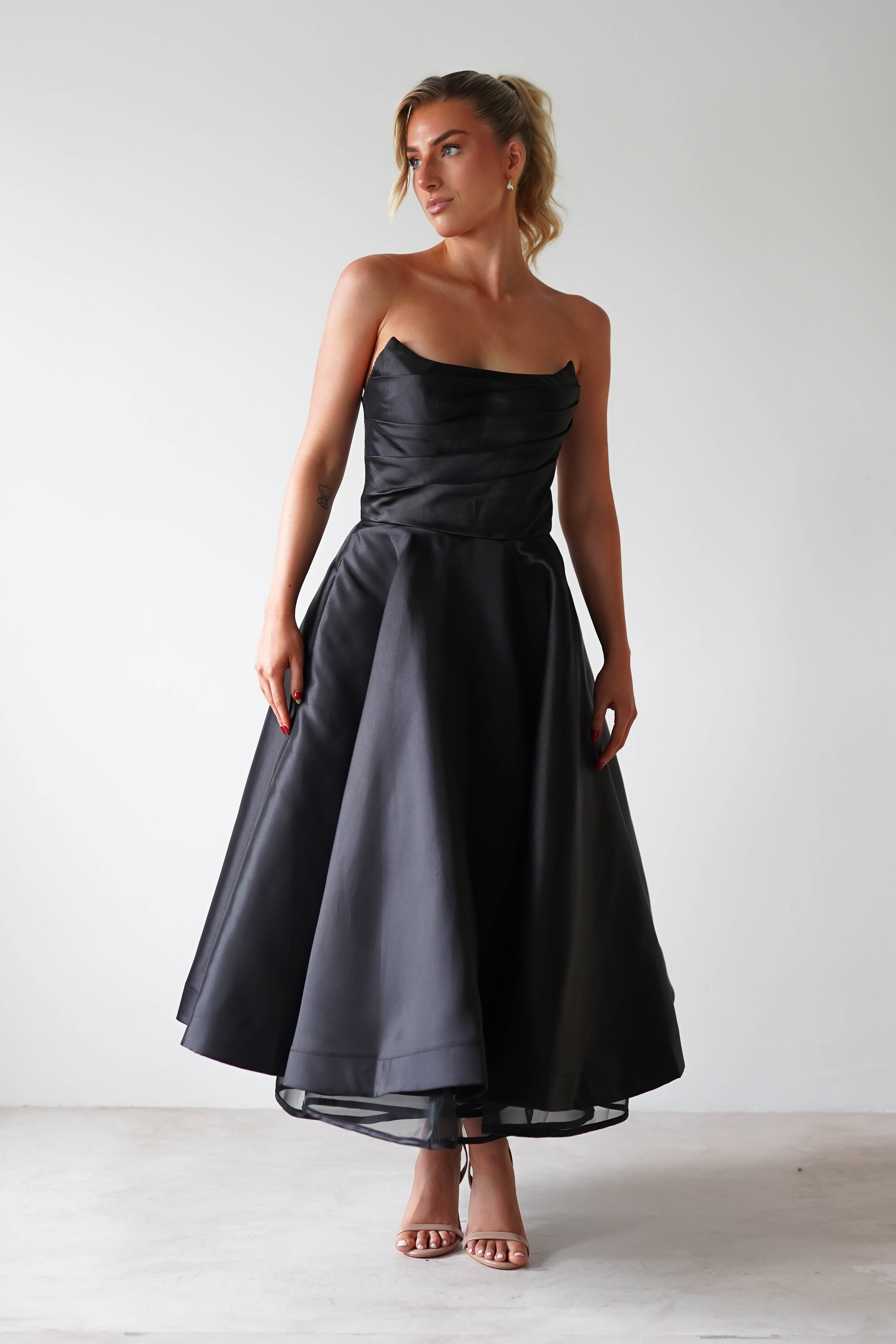 A woman stands gracefully against a plain background, wearing a strapless midi dress crafted from luxurious satin fabric that glistens subtly. The ankle-length black dress features a fitted bodice and flowing skirt, perfect for formal events. Her hair is elegantly styled up, complemented by neutral-toned high-heeled sandals.