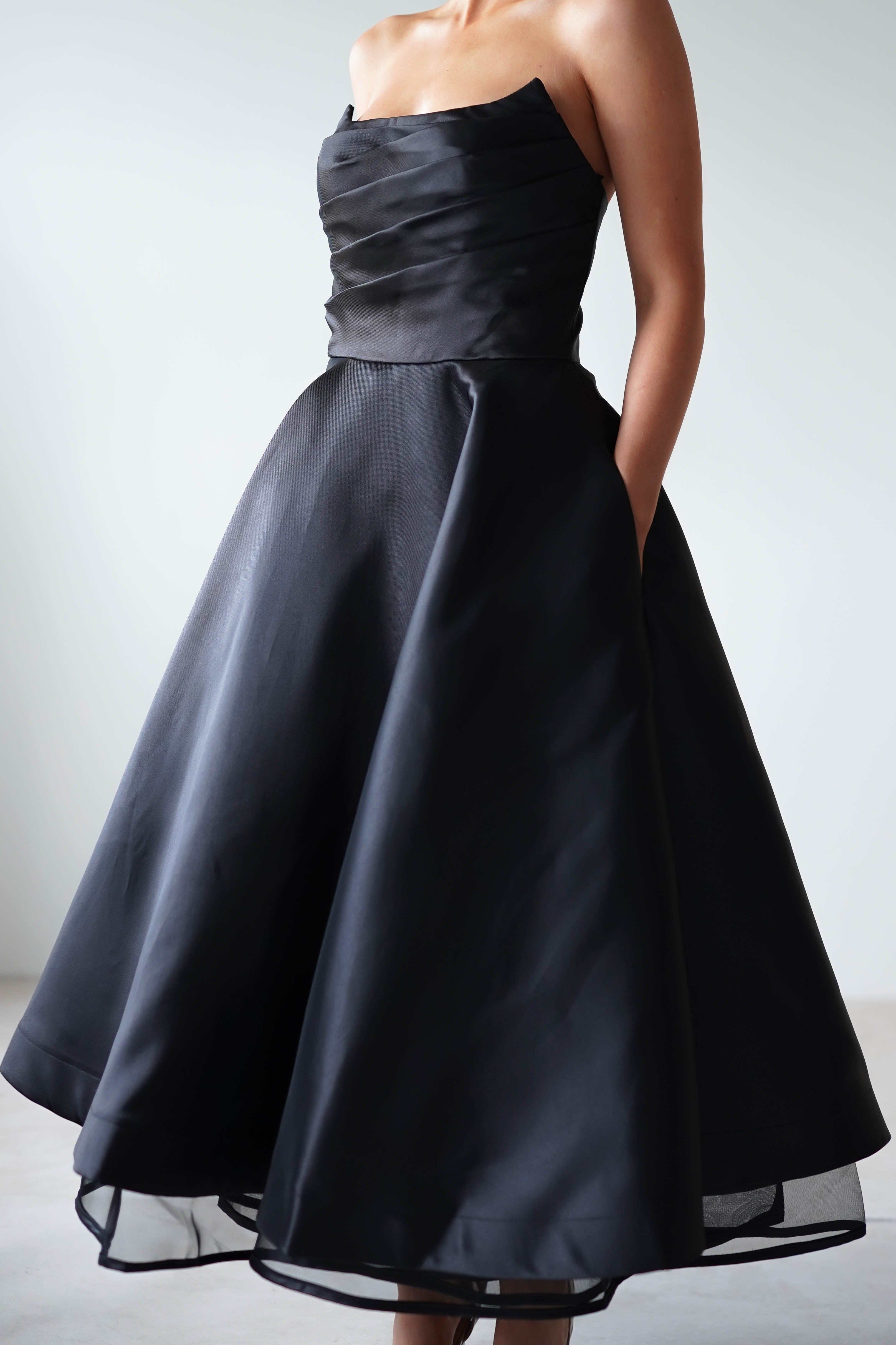 Person wearing an elegant, strapless black evening gown crafted from satin fabric, featuring a fitted bodice and a full, flowing skirt. Their hands are casually placed in the dress pockets, providing a relaxed yet sophisticated look. The background is a simple, muted tone.
