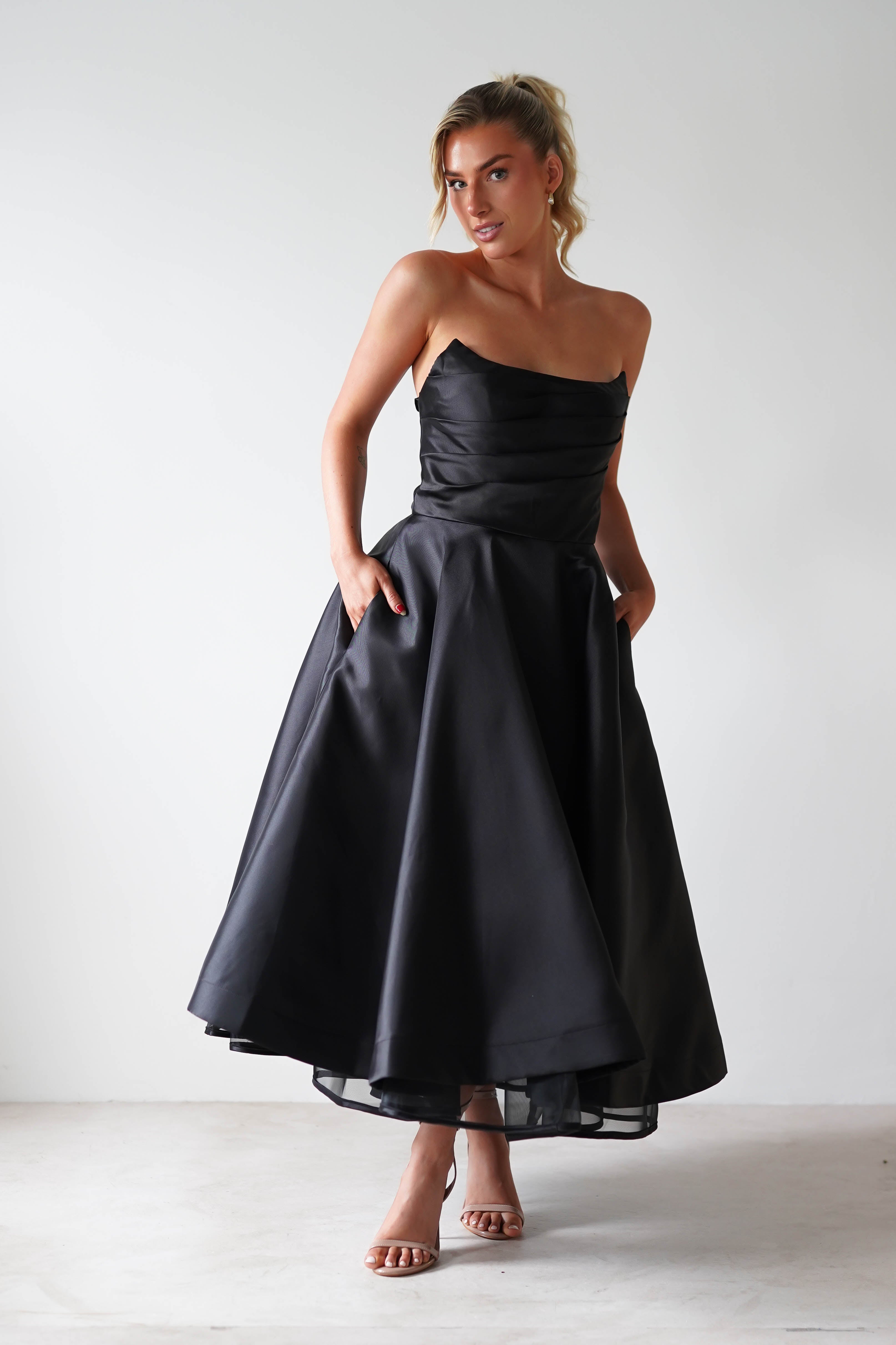 A person in a strapless midi dress made of luxurious satin fabric stands against a neutral background, looking to the side with hands in the dress pockets. They wear beige high heels, and their hair is styled up, exuding elegance and poise—perfect for formal events.