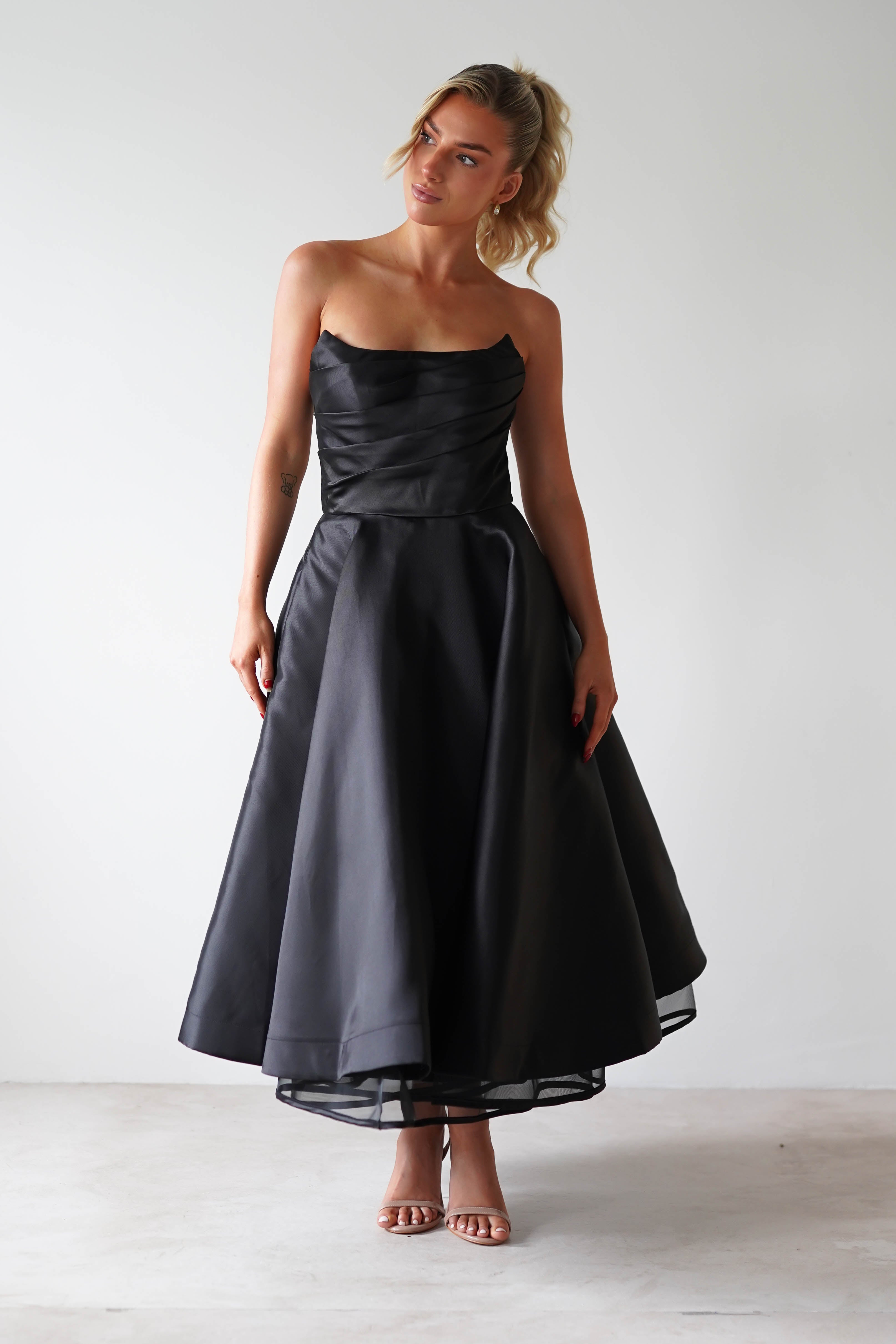 A woman in a strapless black satin midi dress stands against a plain background. Perfect for formal events, the dress features a fitted bodice and flowing skirt. Her blonde hair is styled up, complemented by simple earrings.