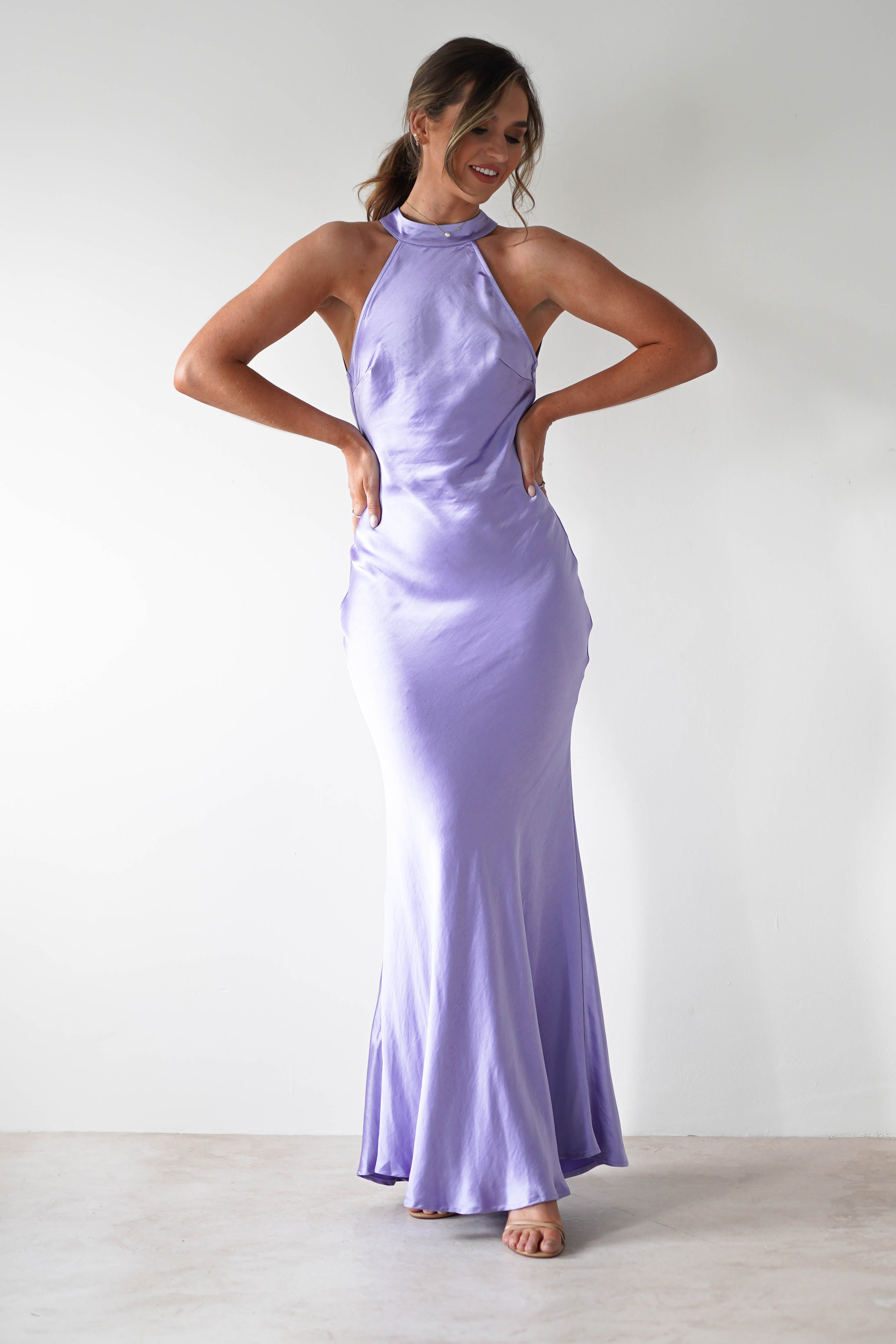 A woman stands against a plain white wall, smiling and posing with her hands on her hips. She wears a long, light purple halterneck maxi dress made from satin material and open-toe heels.