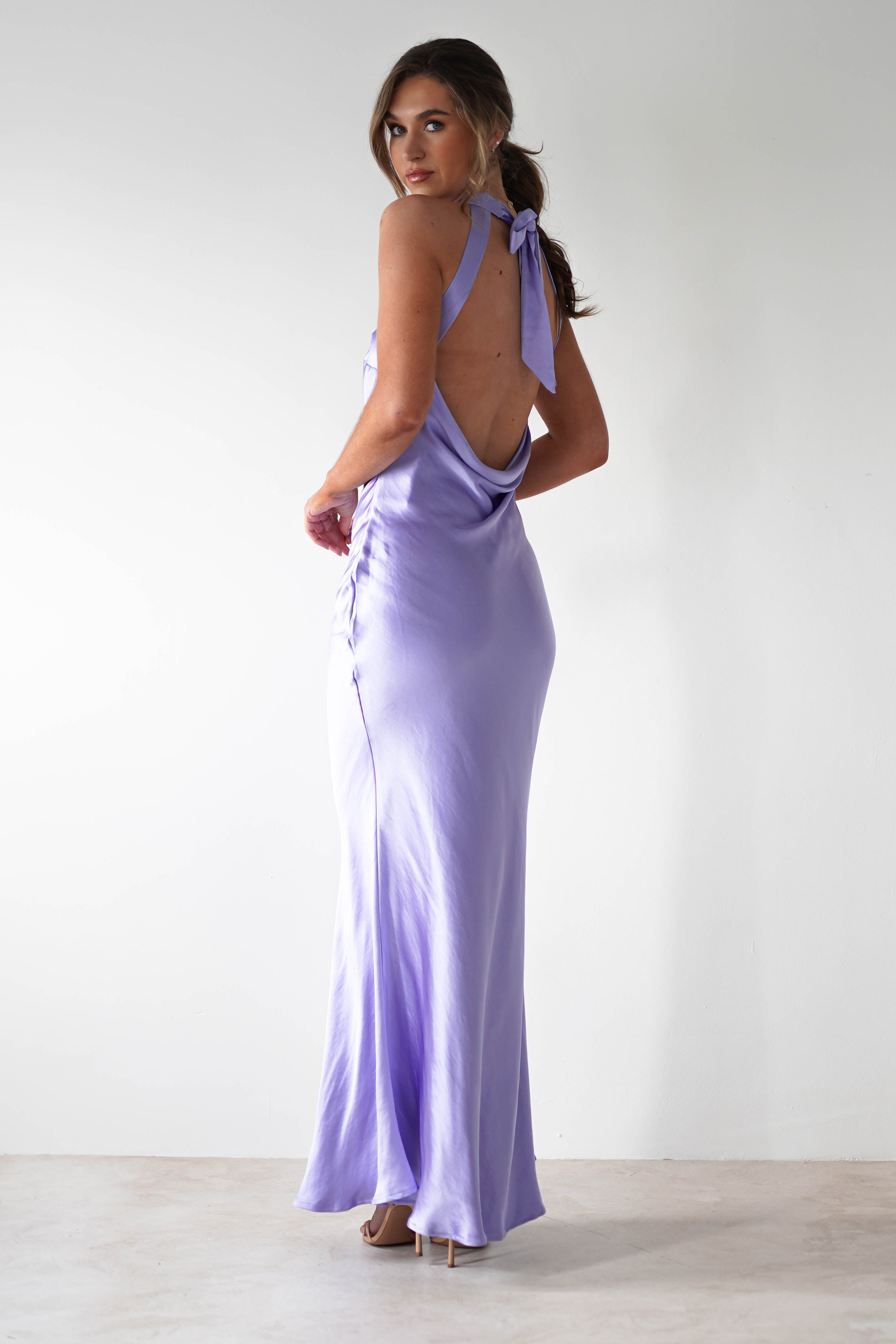 A woman stands in a minimalist room wearing a halterneck maxi dress in lavender satin material with an open-back detail and a bow tie at the neck. She faces away but looks over her shoulder, hair in a loose ponytail and high heels on her feet.
