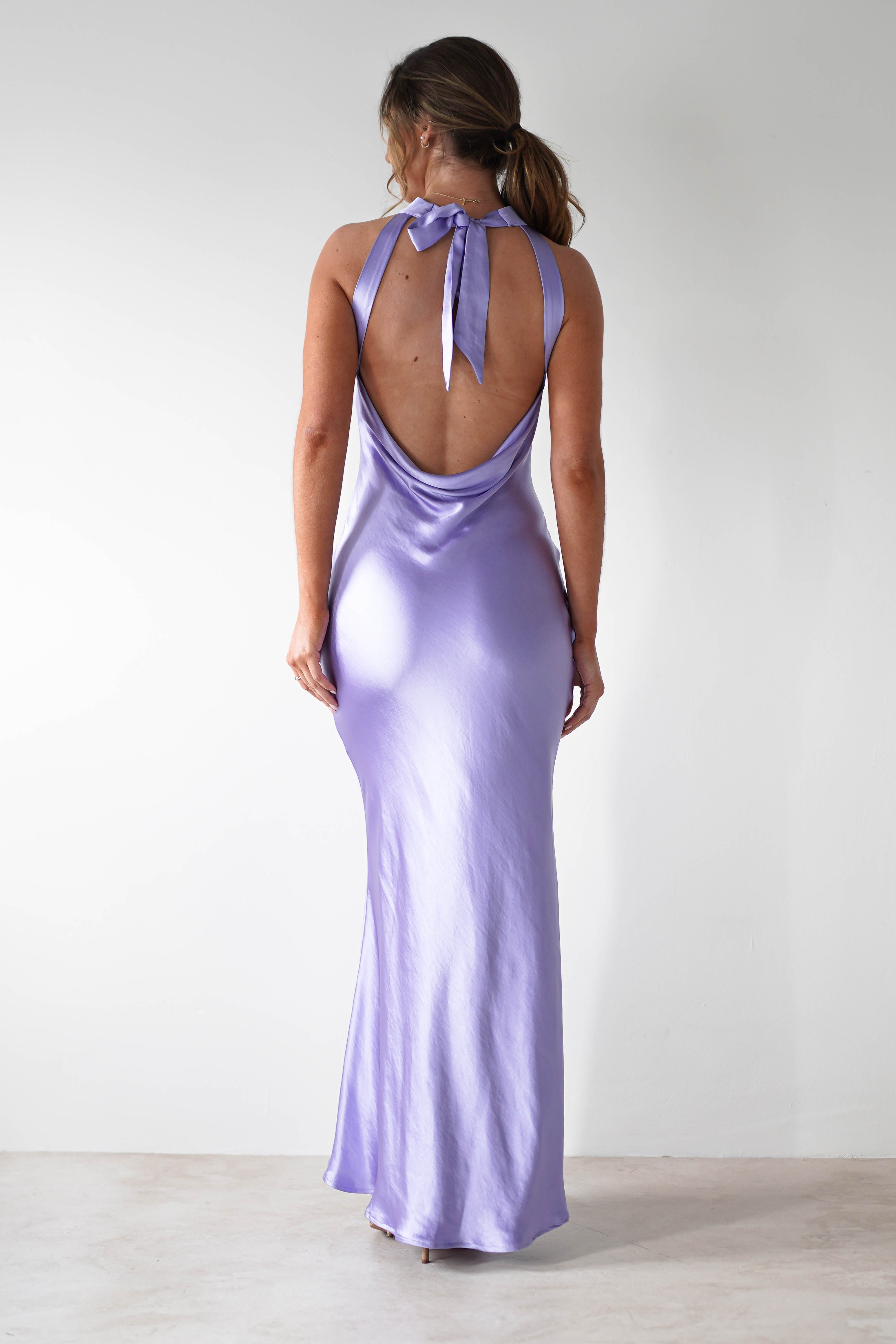 A woman stands facing away, wearing a shiny lavender satin material halterneck maxi dress with an open-back detail and a bow at the neck. Her hair is pulled back, posed against a plain white wall.