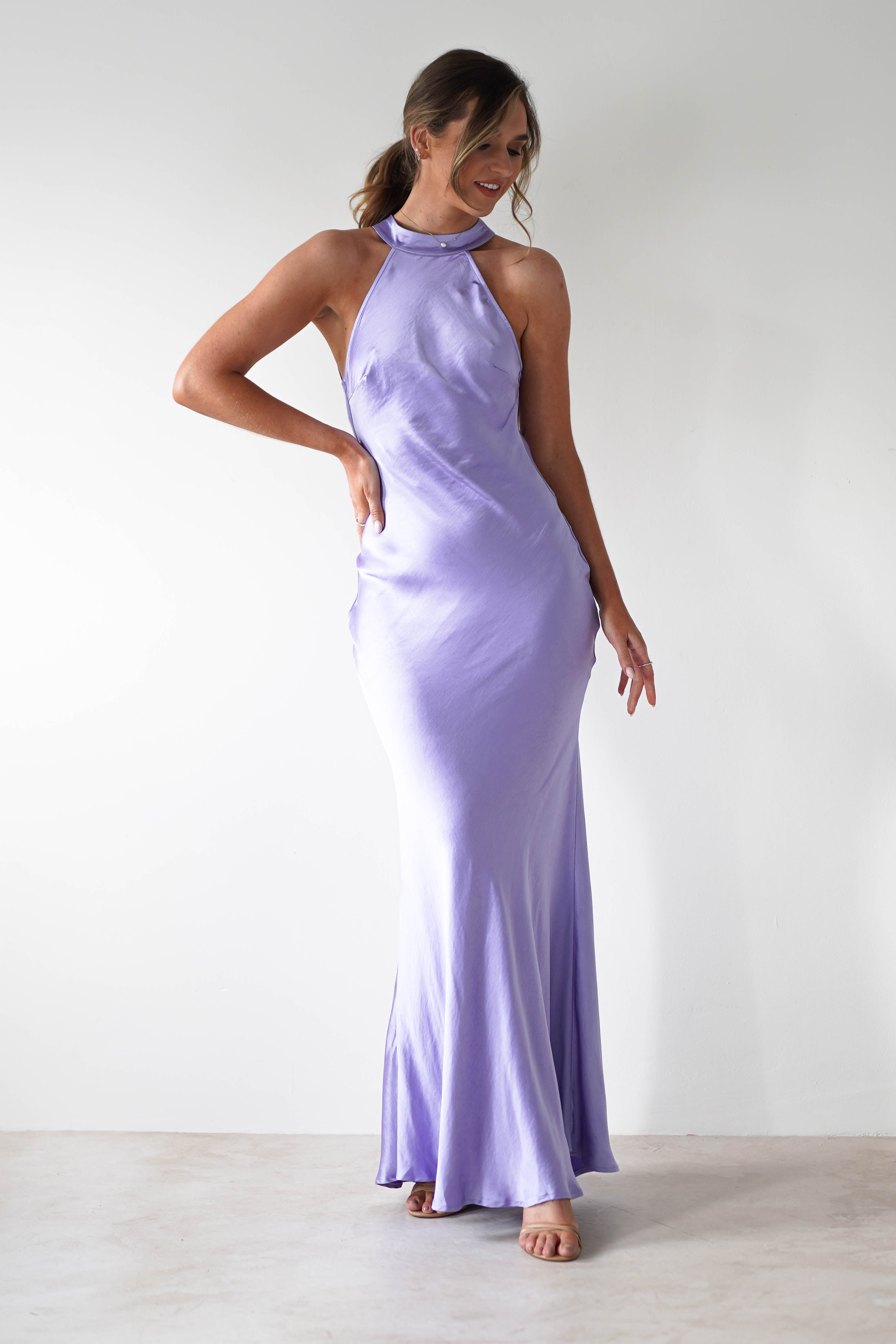 Woman standing against a plain white wall, wearing a lilac satin halterneck maxi dress with an elegant open-back detail. She has her hand on her hip, is smiling, and wears her hair tied back in a low ponytail.