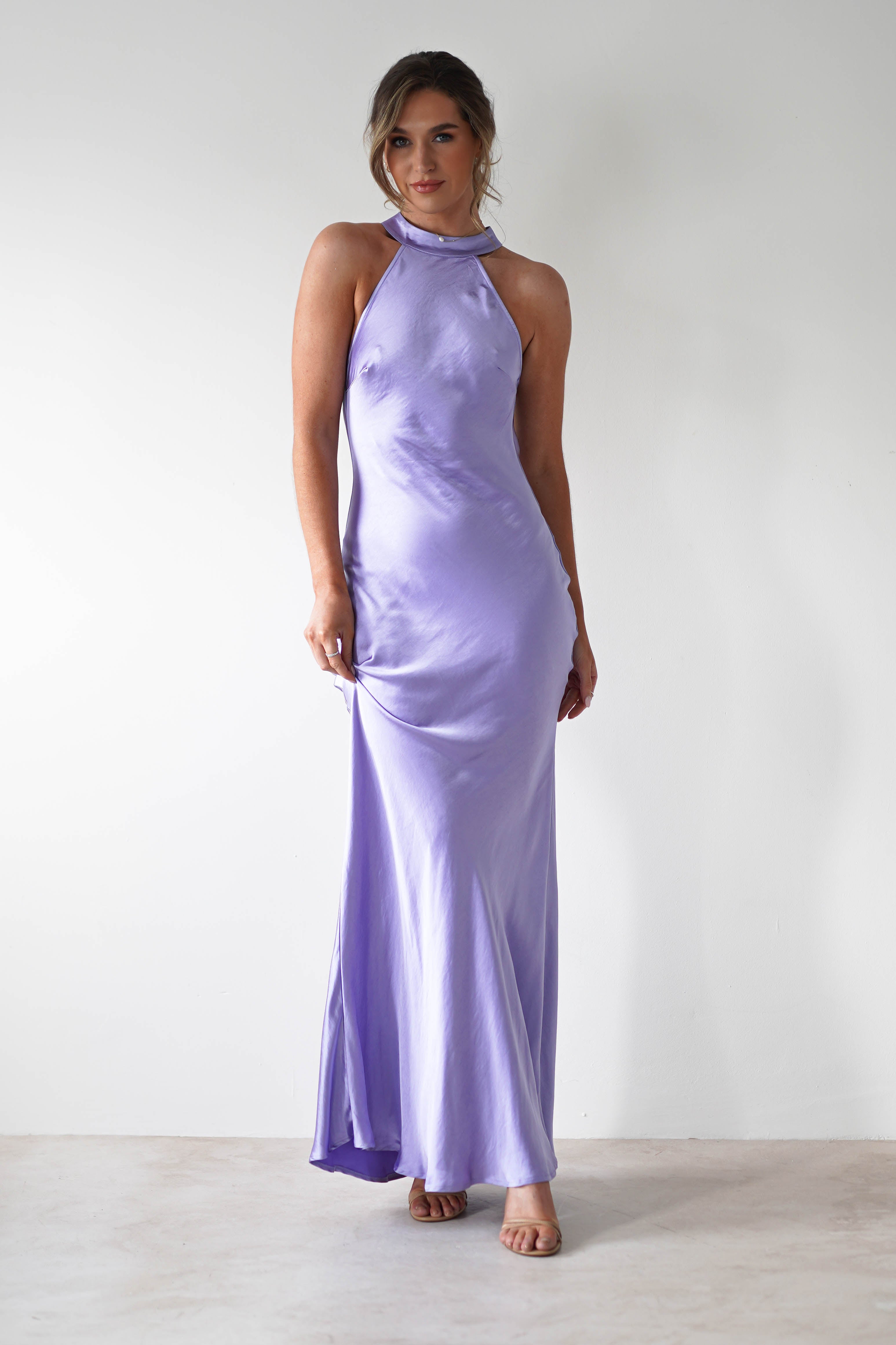 A woman stands against a plain white wall wearing a halterneck maxi dress in lilac, crafted from thick satin material. The floor-length dress features open shoulders, while her light brown hair is styled up and she pairs the look with open-toe heels.