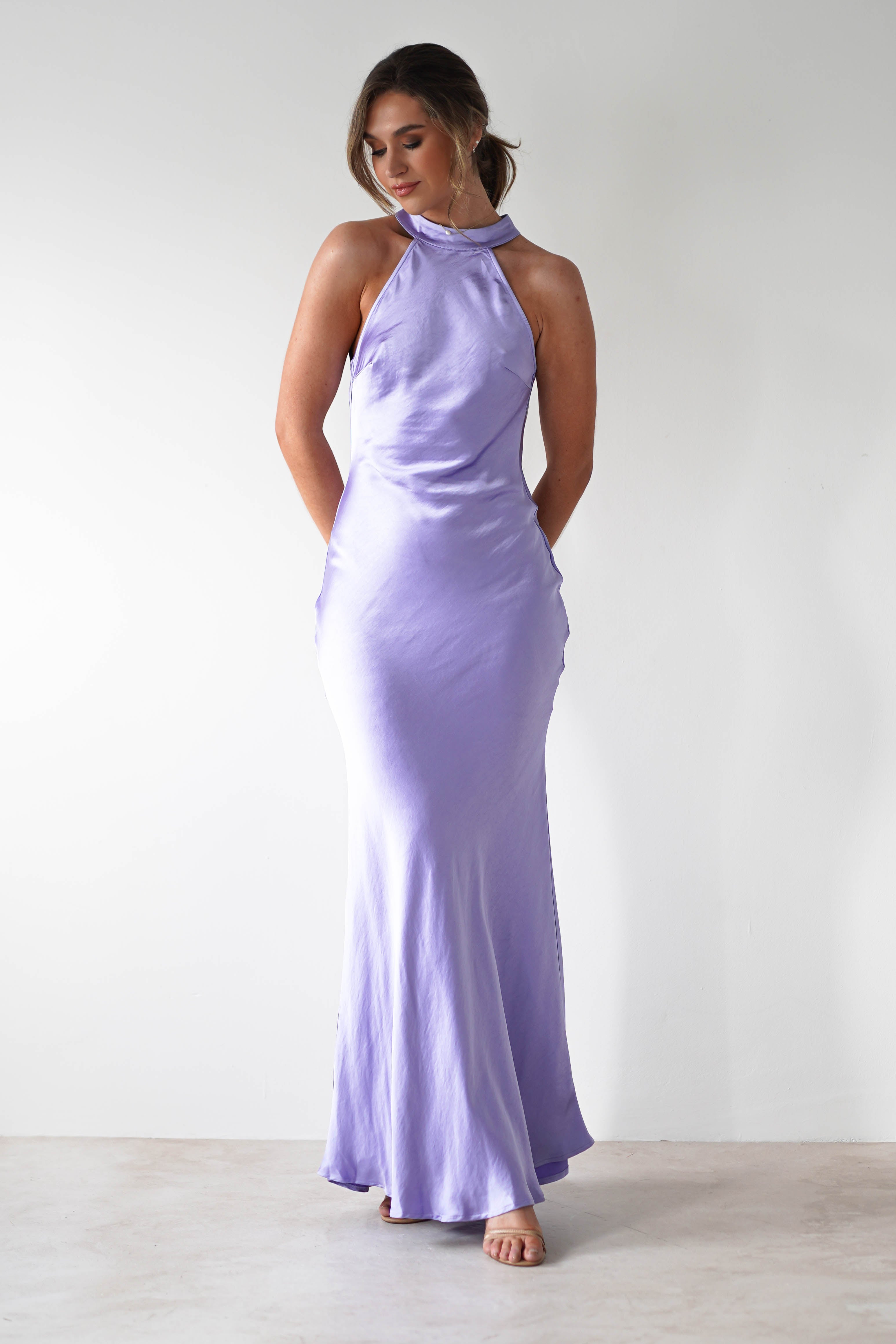 A woman stands against a plain white background wearing a pale lavender halterneck maxi dress in thick satin material, featuring an open-back detail and subtle train. Her hair is styled in a loose updo as she stands barefoot, looking slightly downward.