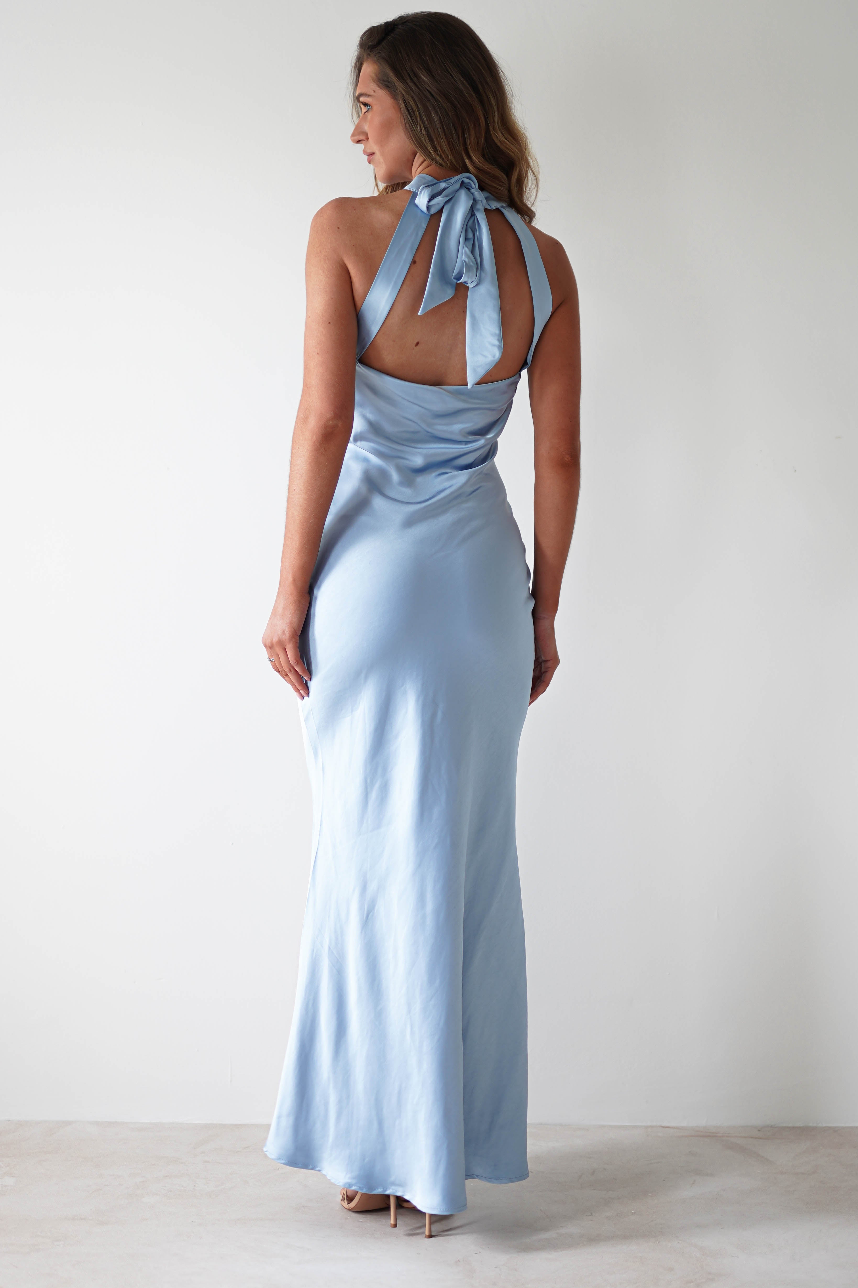 A woman with long hair stands against a plain white background, showcasing an elegant halterneck maxi dress. The light blue satin material drapes beautifully, highlighting the open-back detail as it ties gracefully at the neck.