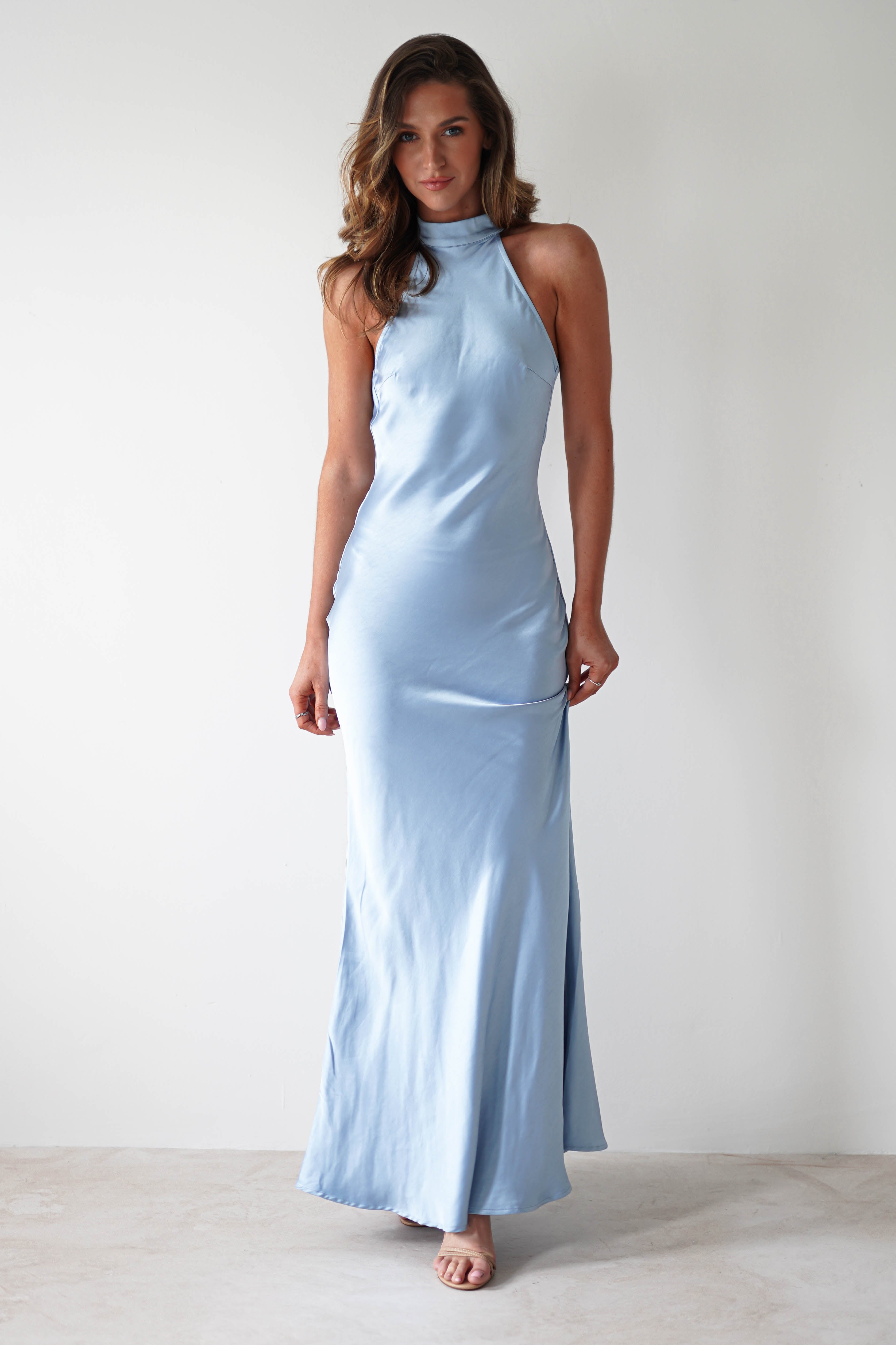 A woman stands against a plain white background, elegantly donning a satin halterneck maxi dress in light blue. The floor-length gown, featuring an open-back detail, complements her long, wavy hair as she smiles warmly at the camera, her left hand gently holding the dress.