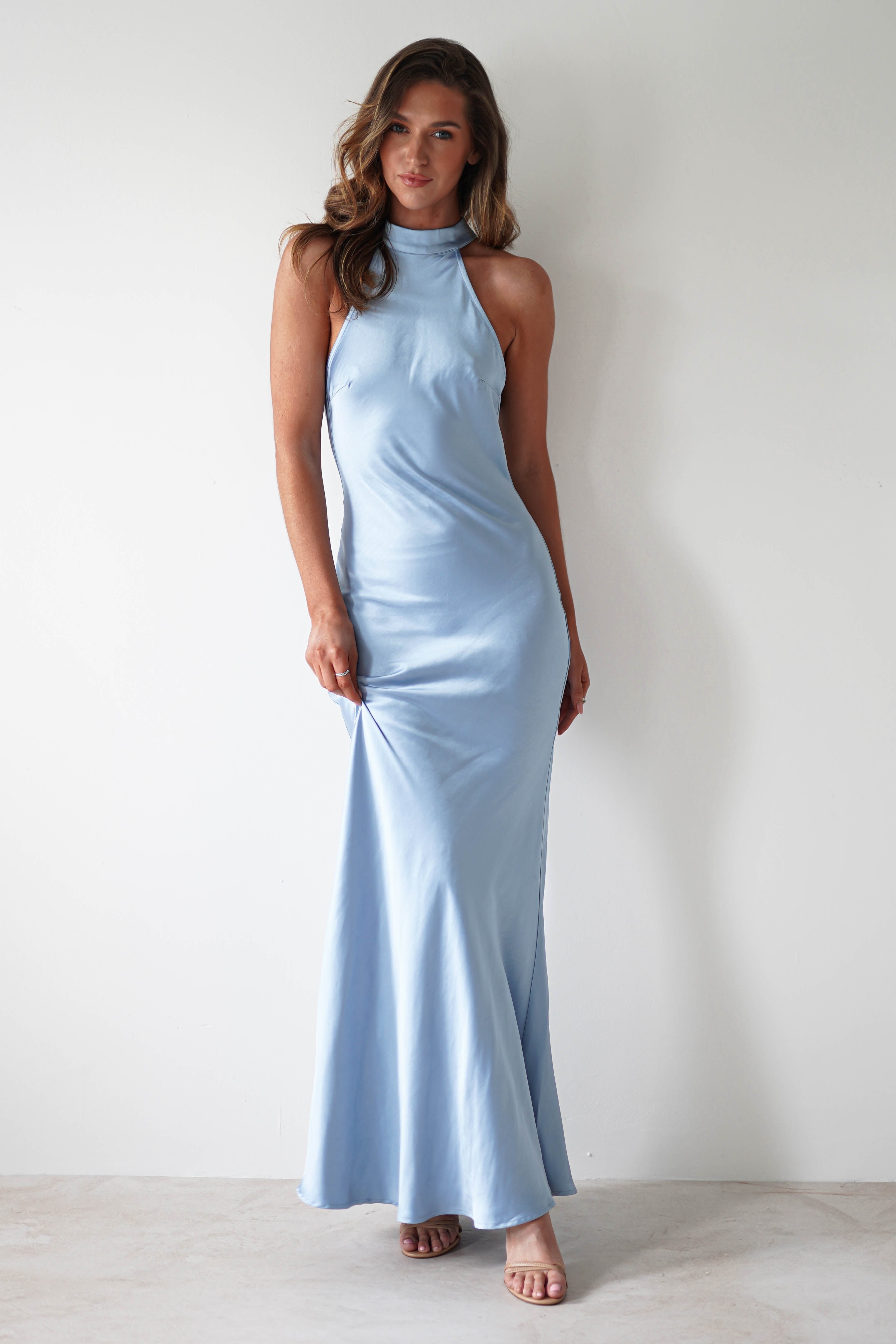 A woman stands elegantly against a plain white background in a floor-length, light blue halterneck maxi dress made of satin material. She wears open-toed heels, her hair styled in loose waves, and her relaxed pose reveals the open-back detail as she gently holds the dress.