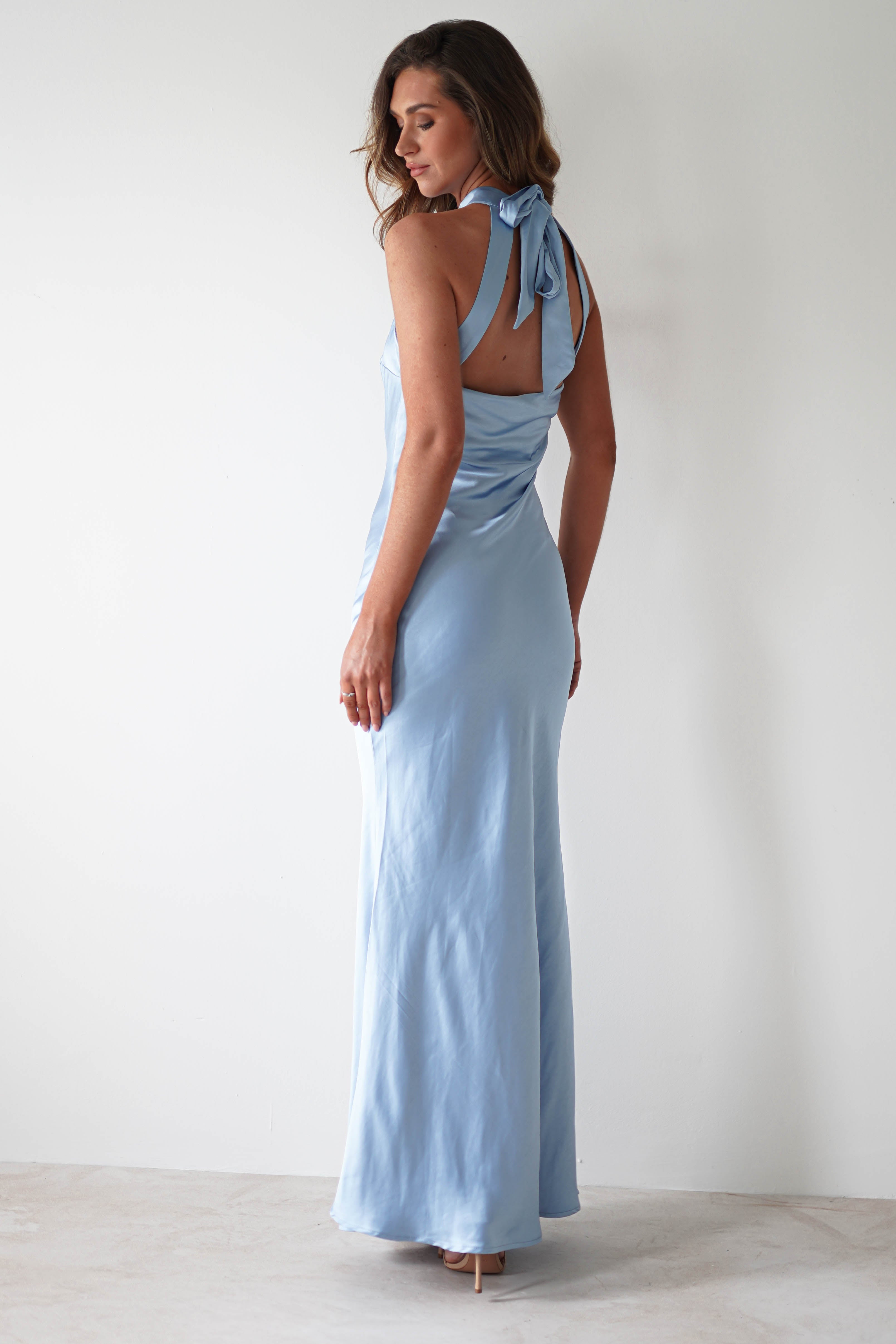 A woman in a silky, light blue halterneck maxi dress stands with her back to the camera, showcasing the stunning open-back detail. The dress features a bow at the neck and flows elegantly to the floor. Her long, wavy hair cascades down against a plain, light-colored wall.