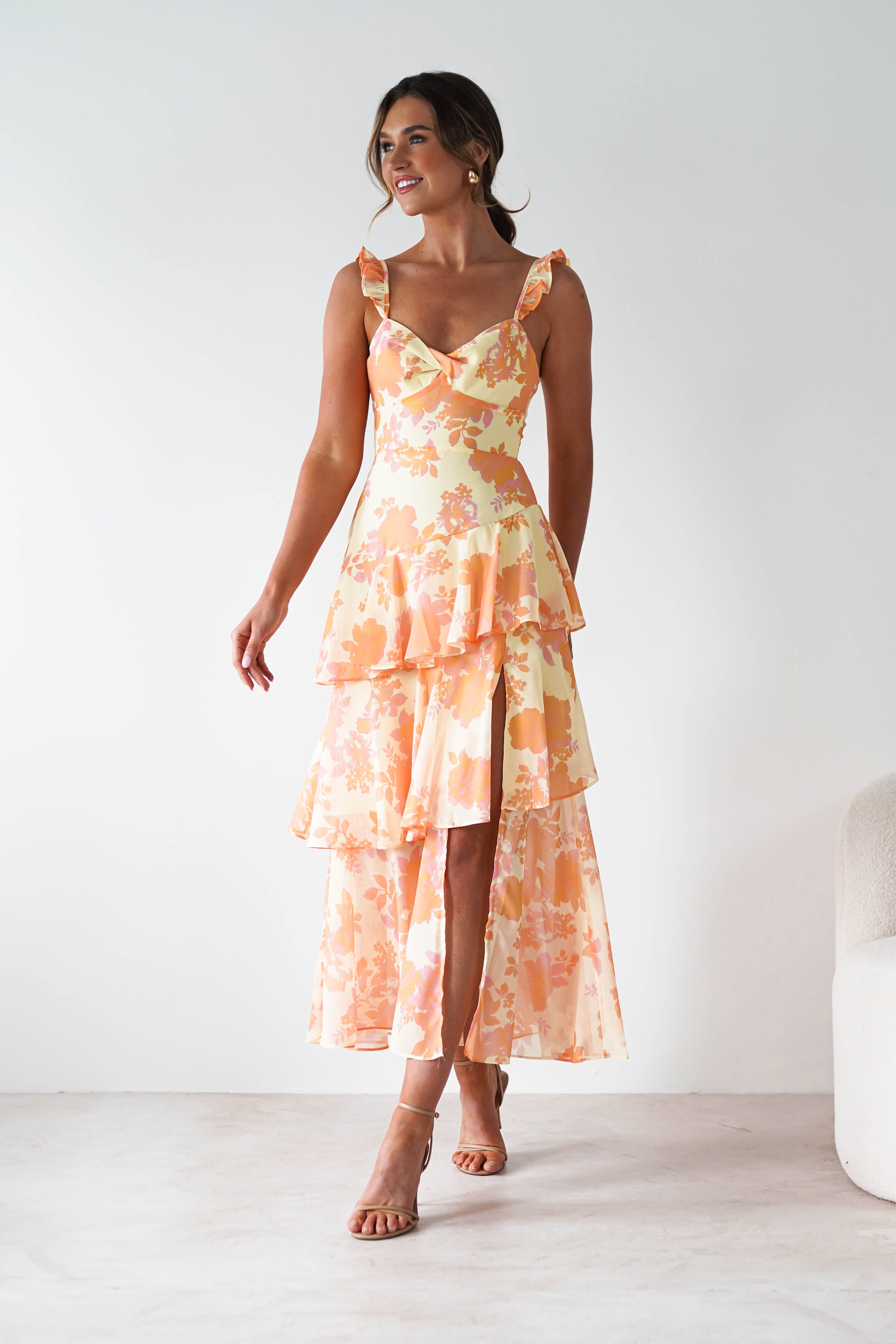 A woman wearing a sleeveless, tiered, peach and orange floral midaxi dress with ruffle straps stands indoors on a light floor, smiling and looking to the side. She wears strappy heels and has her hair styled up—perfect as a wedding guest dress.