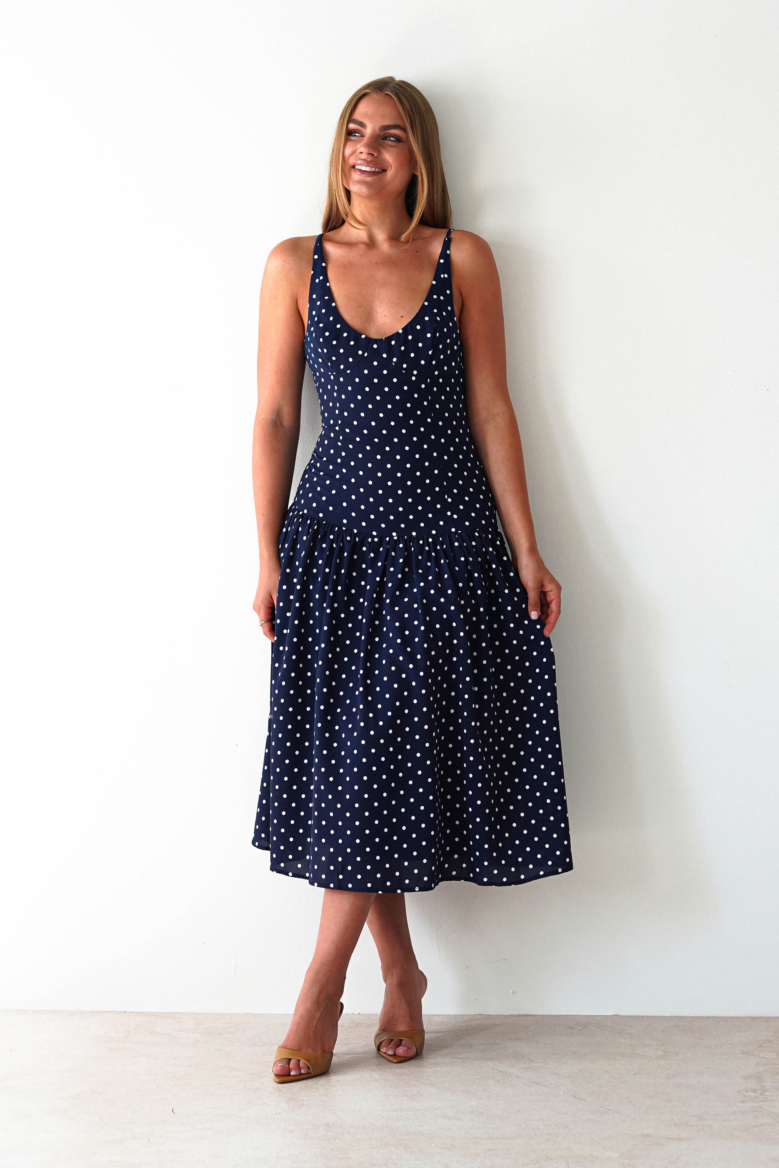 Woman wearing a navy blue sleeveless midi dress with white polka dots and a scoop neckline, paired with open-toe tan heels.