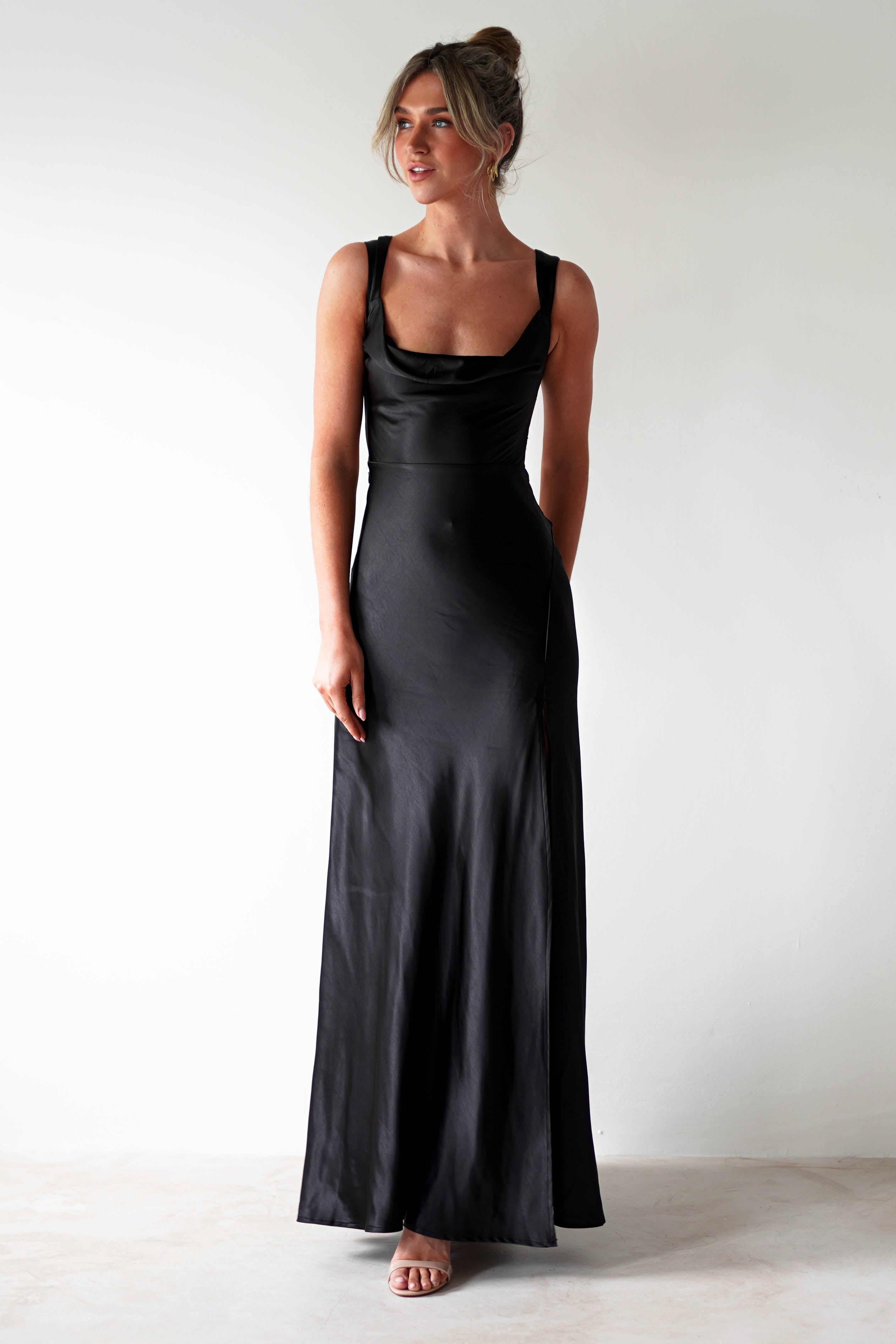 A woman stands against a plain white wall, wearing a long black satin gown with a cowl neckline—perfect for formal occasions. Her hair is styled in a loose updo, and she completes the look with open-toed heels.