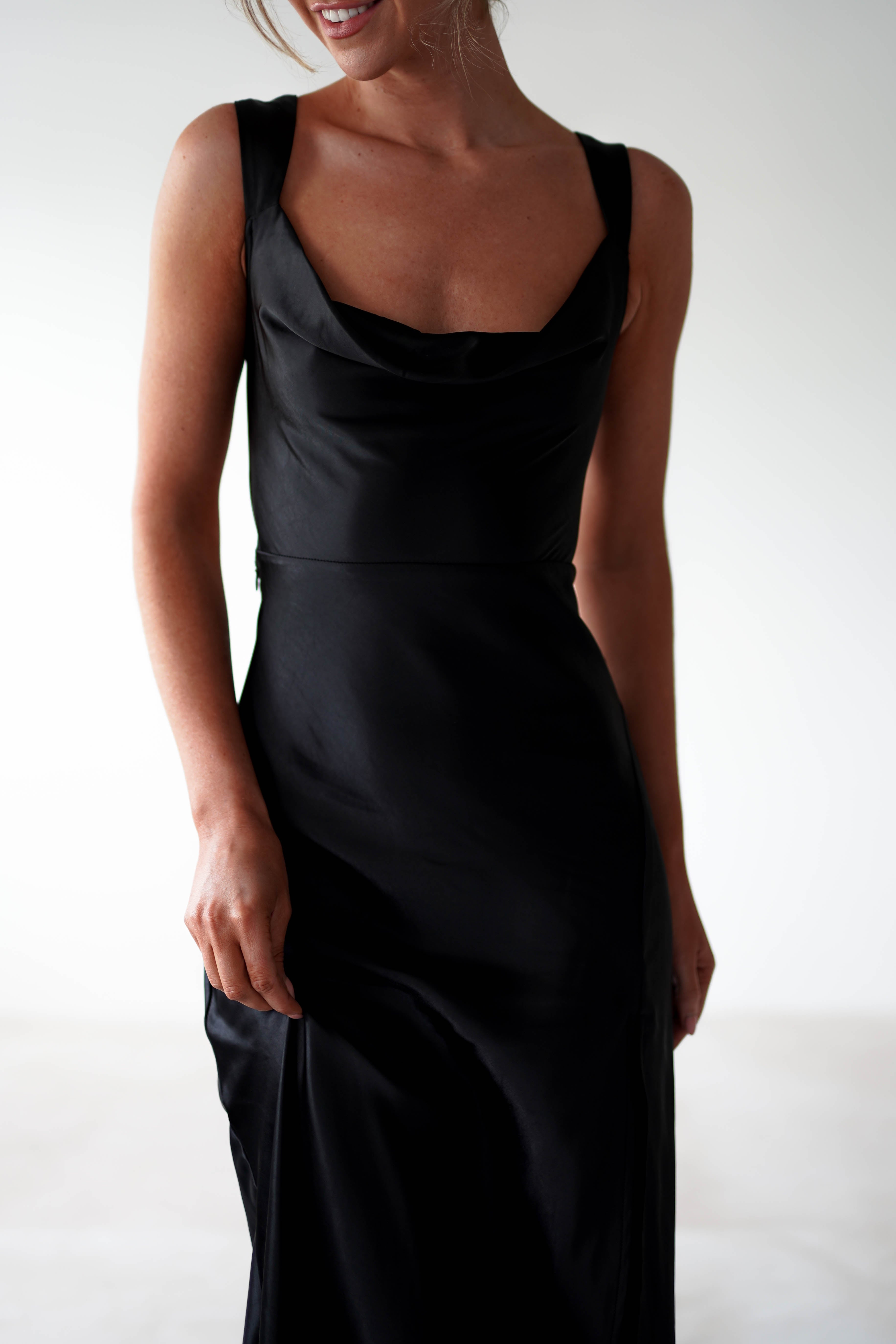 A woman in a sleeveless, fitted black satin gown with a subtle cowl neckline stands against a plain light background. Her head is partially out of frame, and she smiles slightly with her arms relaxed by her sides—a look perfect for formal occasions.