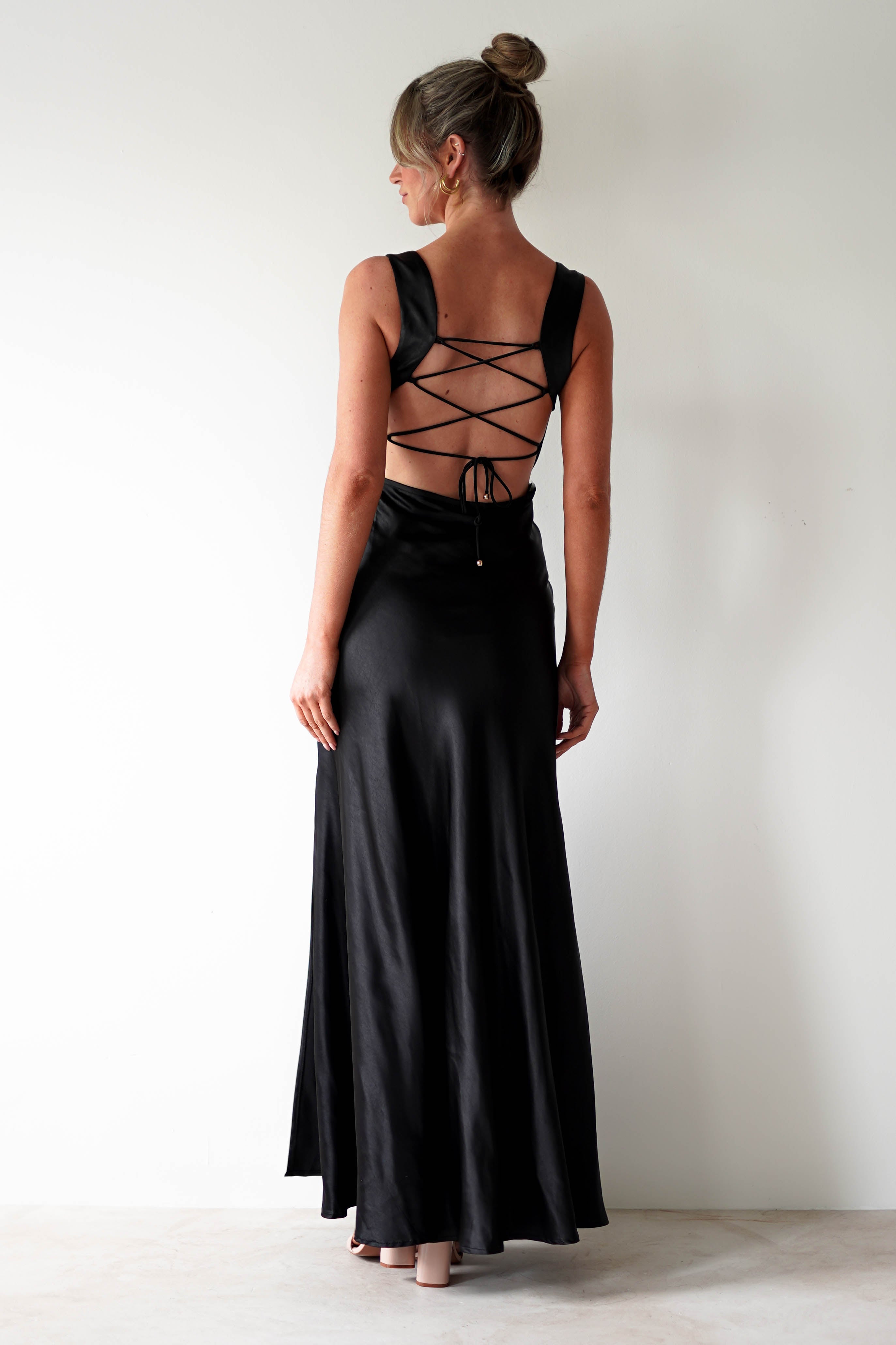 A woman with blonde hair in a bun wears a long, black satin gown with an open back and crisscross lace-up detail—perfect for formal occasions. She stands facing a white wall, looking to the side.