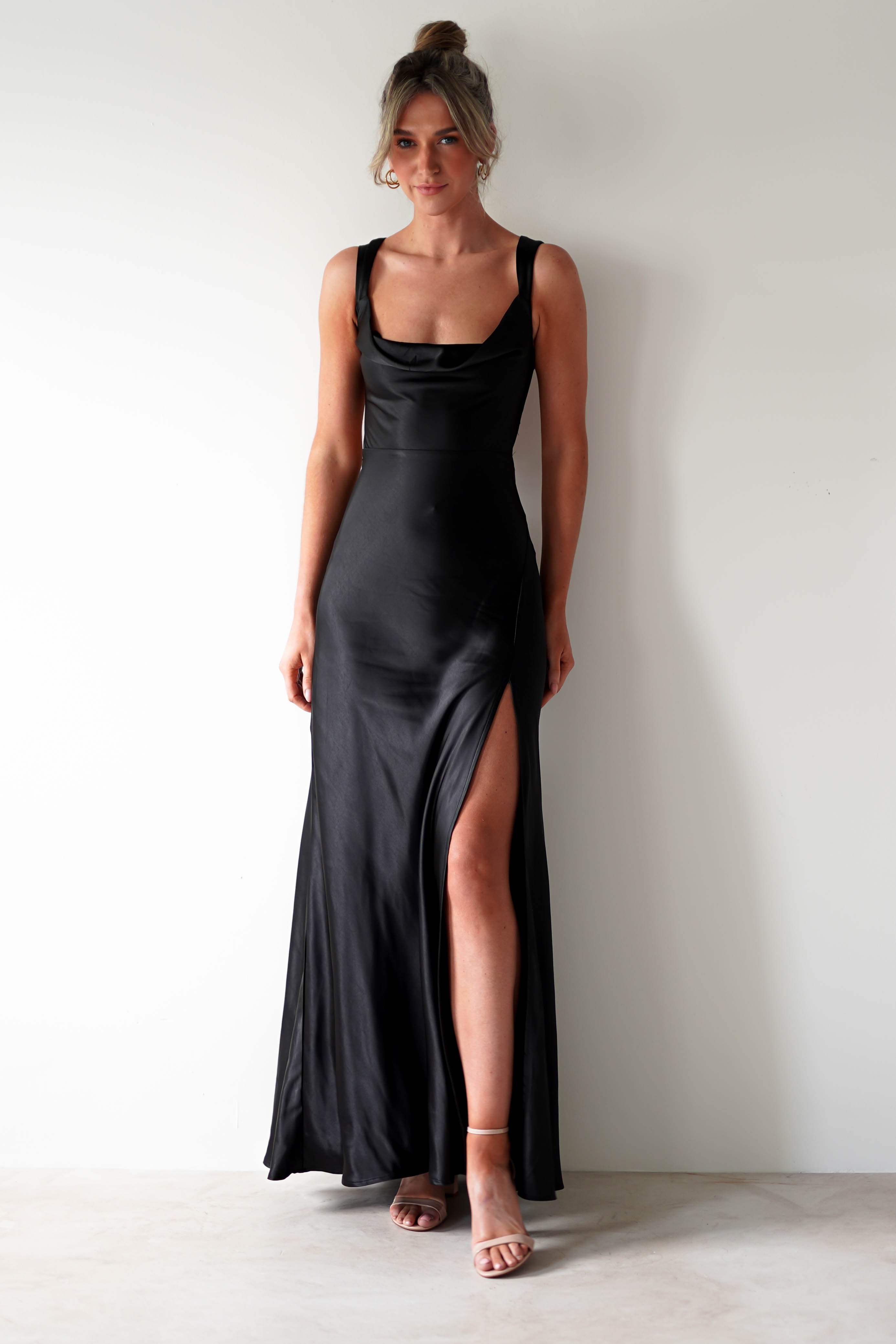A woman stands against a white wall wearing a long black satin gown with a cowl neckline and thigh-high slit, perfect for formal occasions. Her hair is up and she pairs the look with nude high-heeled sandals.