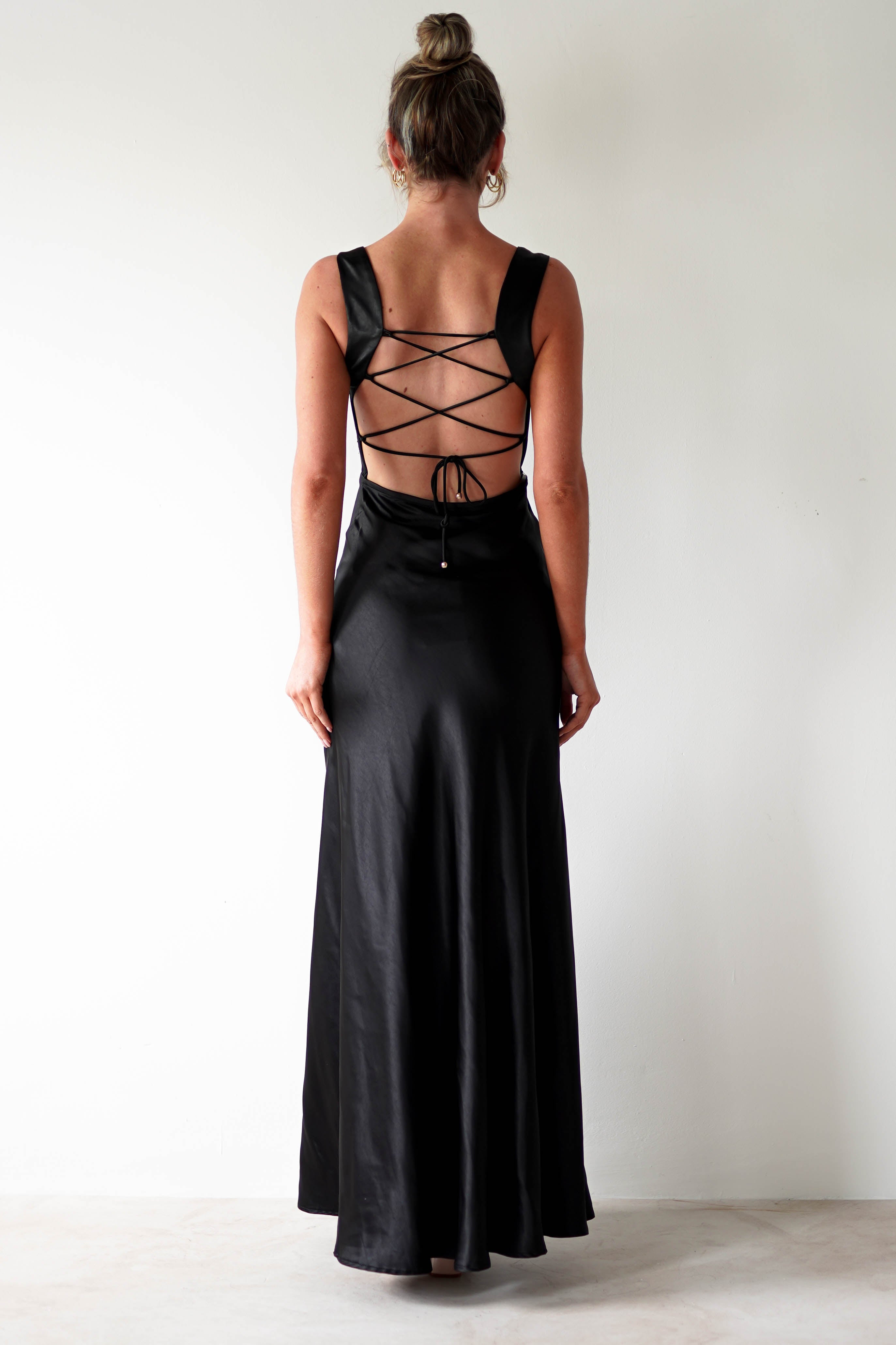 A woman stands facing away, wearing a black satin gown with a cowl neckline and an open back featuring crisscrossed lace-up straps. Her hair is in a bun as she poses against a plain white wall, perfect for formal occasions.